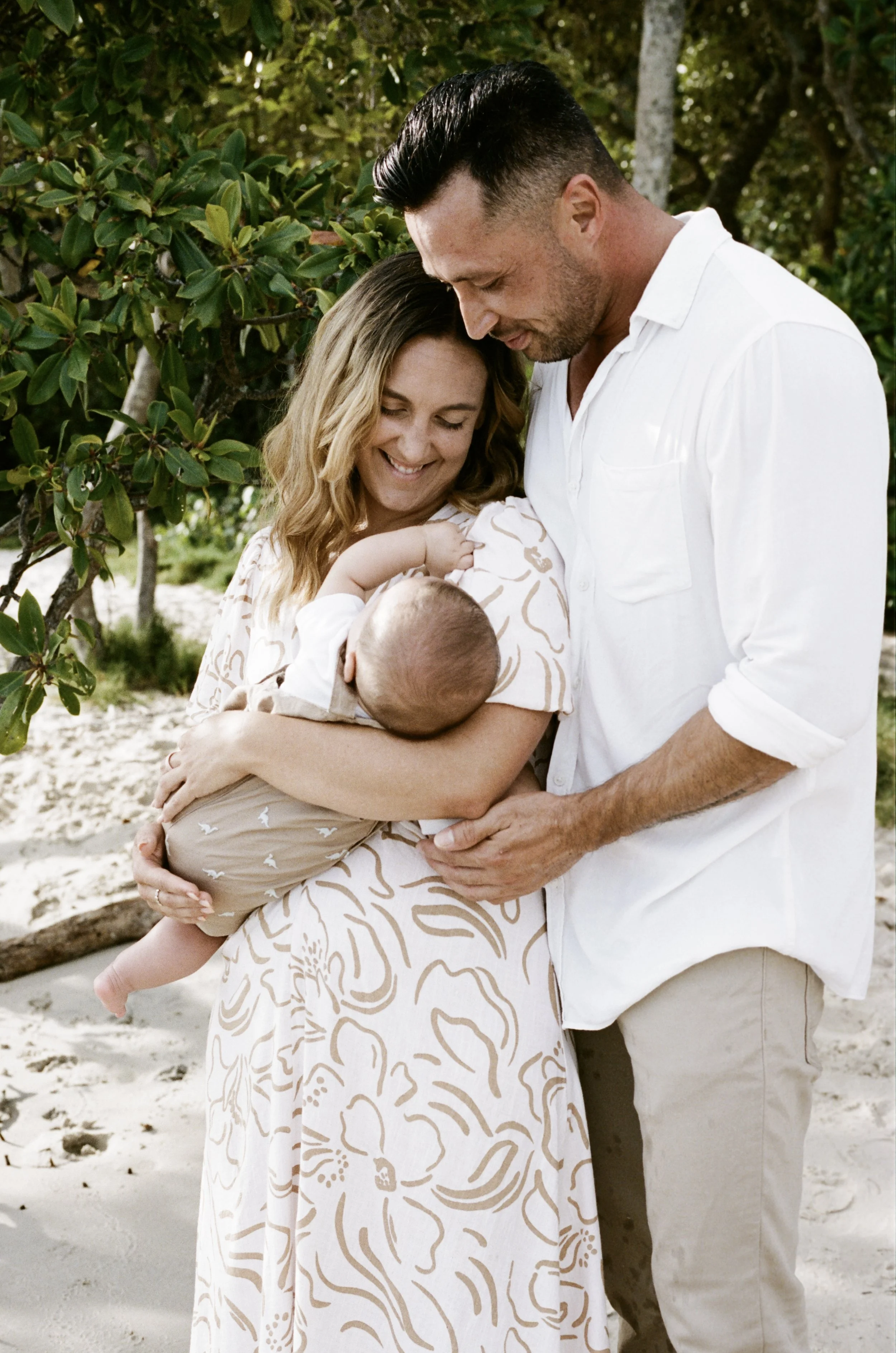 A smiling woman with wavy hair holding a baby, standing with a man on a beach, surrounded by trees and sand.