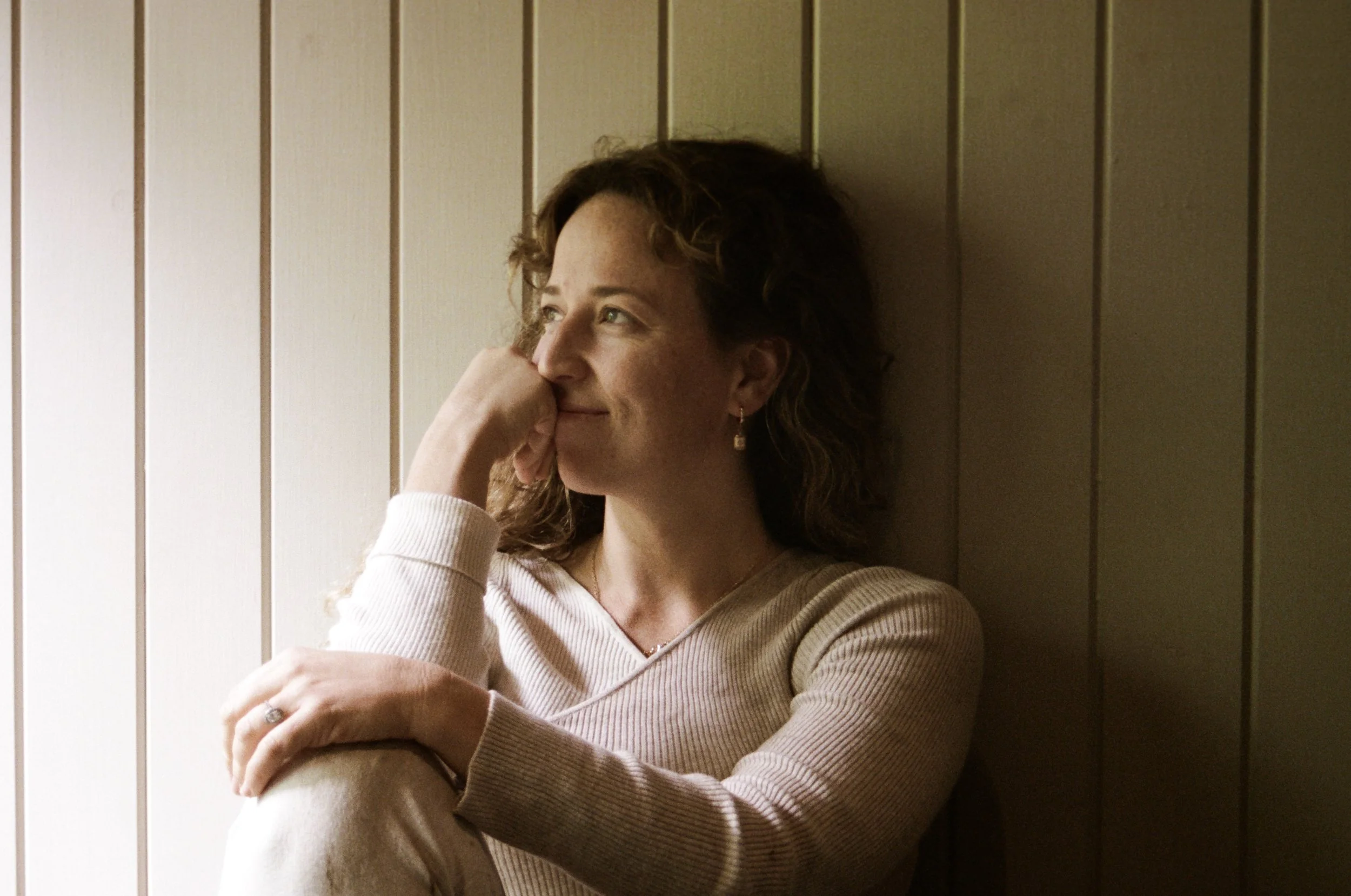 A woman with light brown, curly hair leaning against a wooden wall, wearing a beige V-neck sweater, looking to her right.