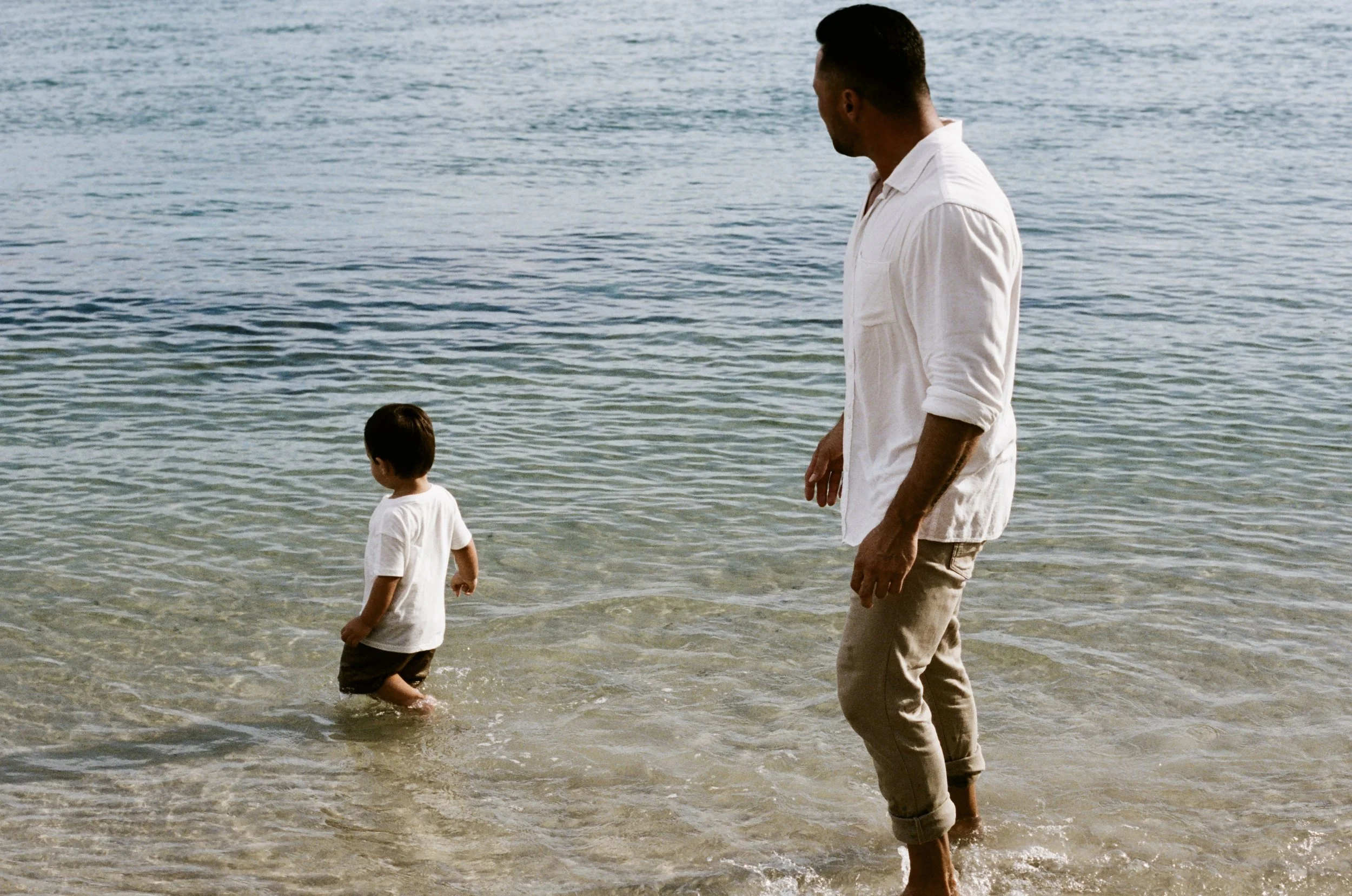 A man and two children walking in shallow water at the beach, with the ocean in the background.