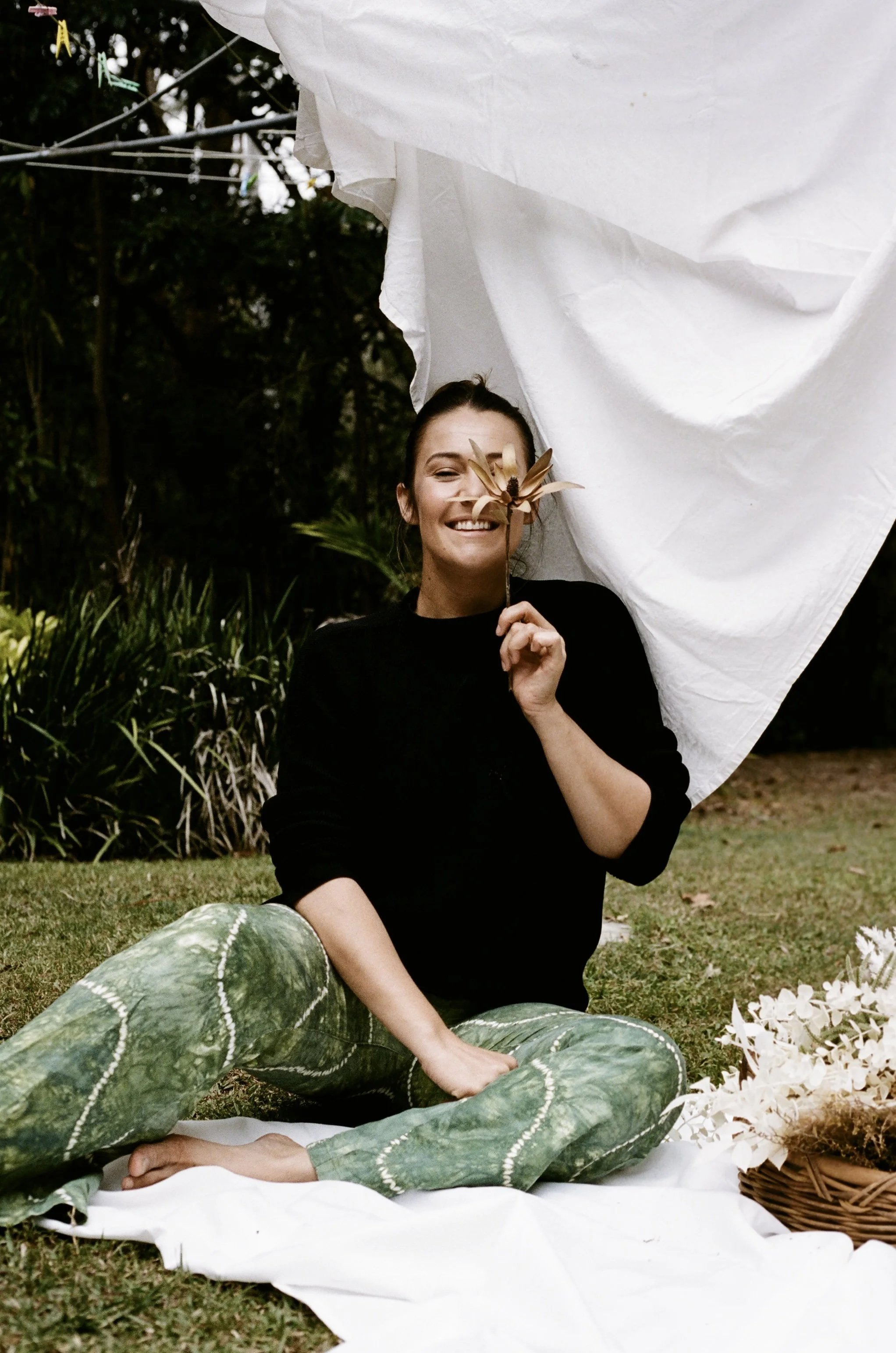 A woman sitting outdoors on a white cloth with a pumpkin in front of her and a basket of white flowers nearby. She is smiling, holding a flower, and sitting against a backdrop of trees and greenery.