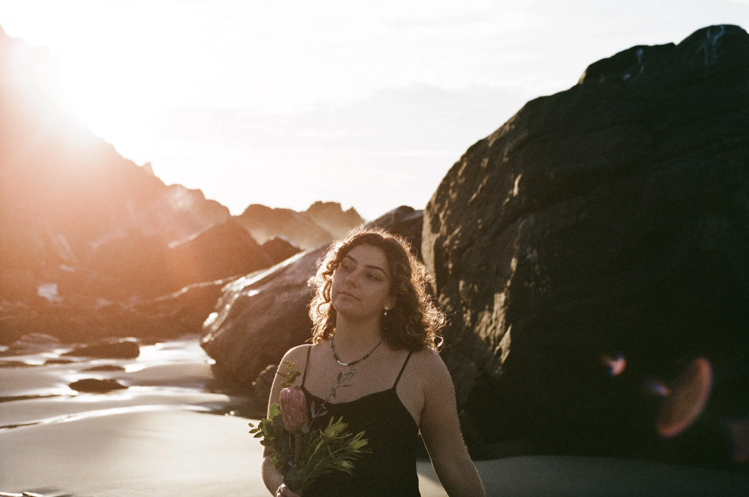 A woman with curly hair holding a bouquet of flowers standing on a beach at sunset with large rocks in the background.