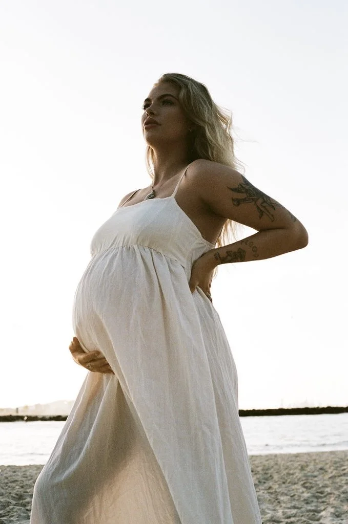 A pregnant woman with tattoos on her arm, dressed in a flowing white dress, standing on a beach at sunset.