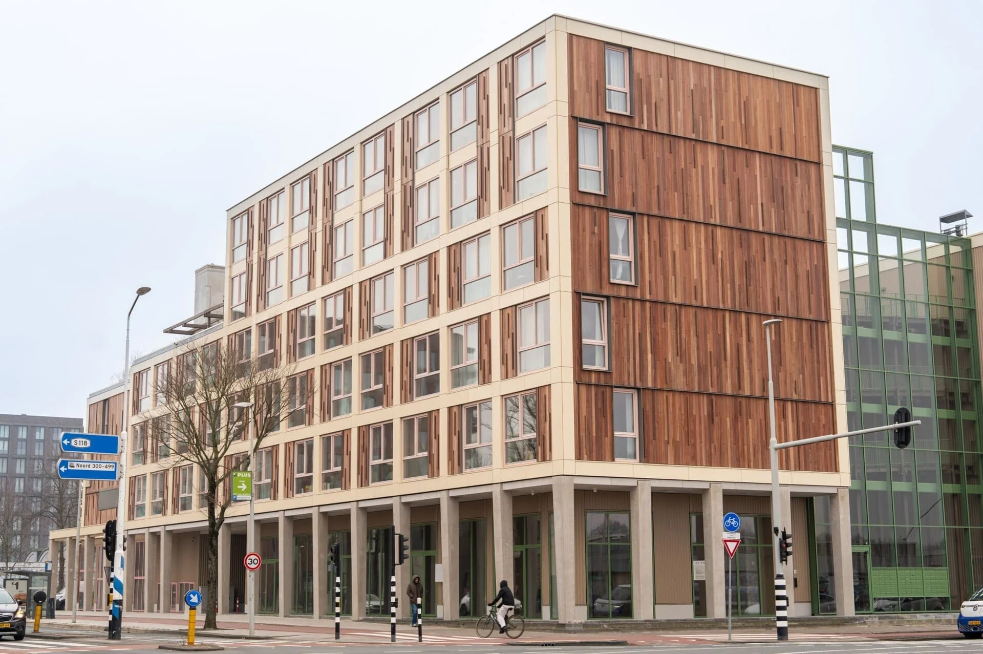 Modern multi-story building with a wooden facade and large windows, situated in an urban area with street signs, traffic lights, and pedestrians.