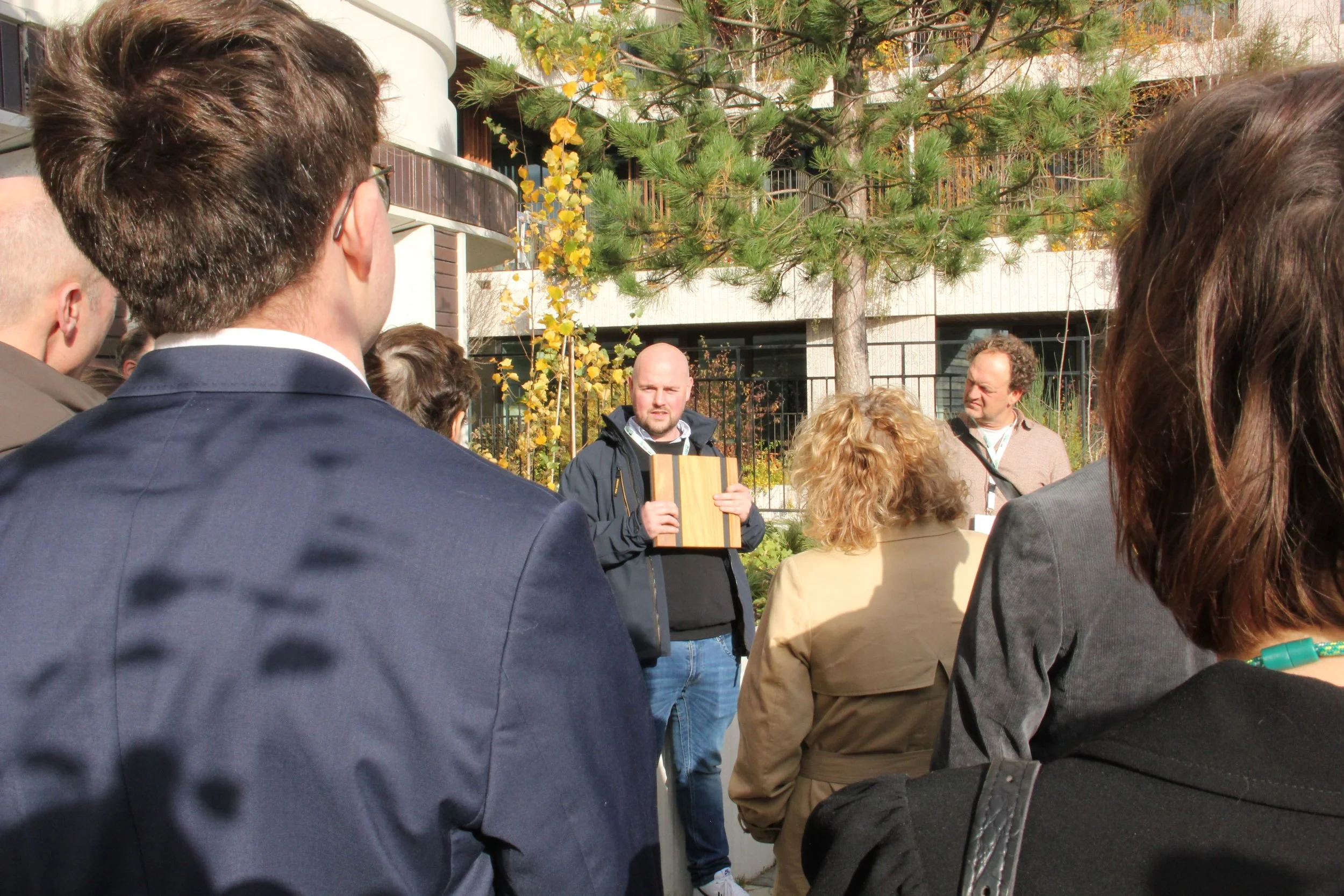 A group of people gathered outdoors, listening to a man speaking and holding a book or binder. The scene includes trees and a building in the background.