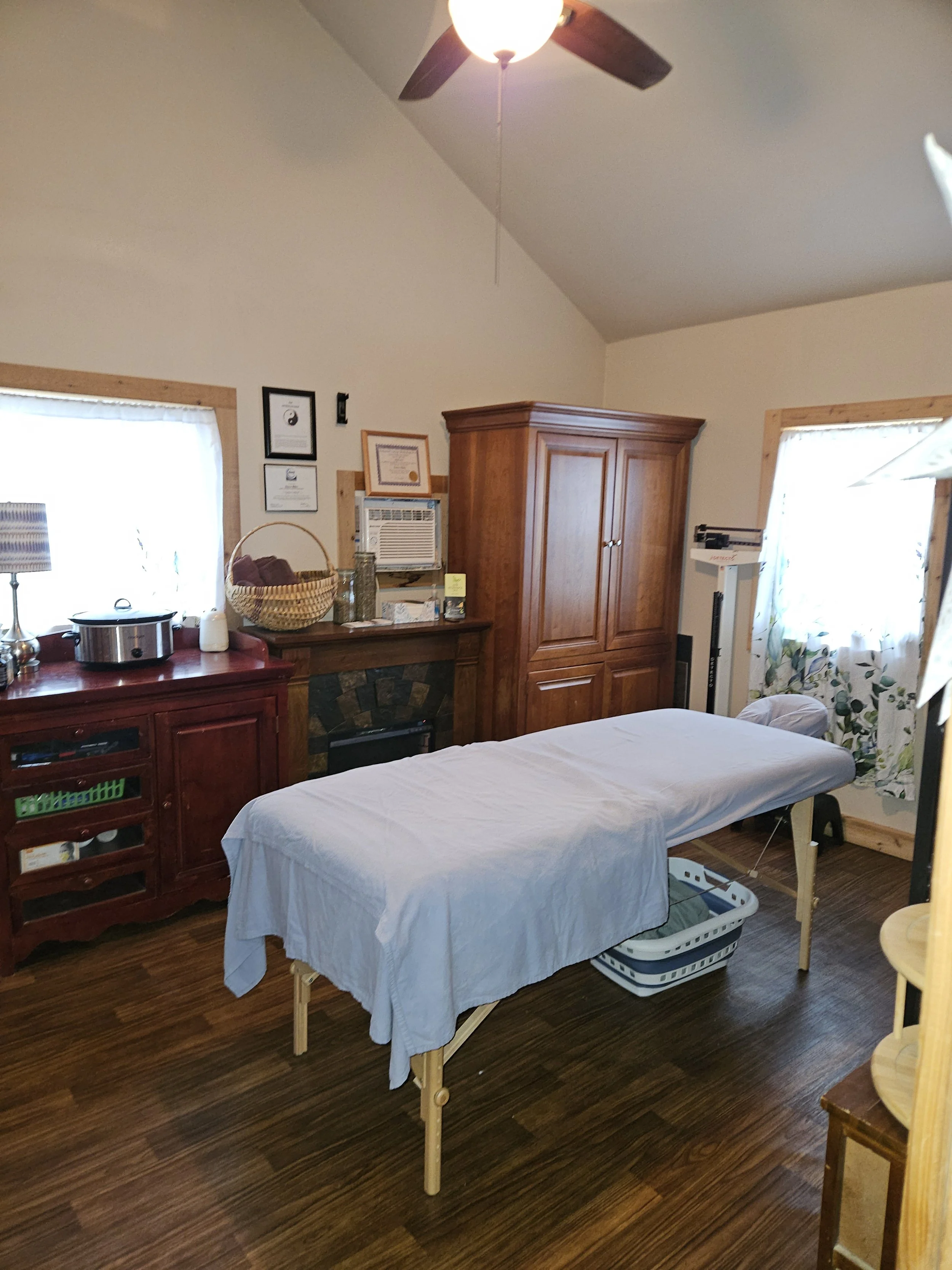 A massage therapy room with a massage table covered in a white sheet, a wooden cabinet, a red sideboard, and a heater. There are two windows with white curtains, and framed certificates on the wall.