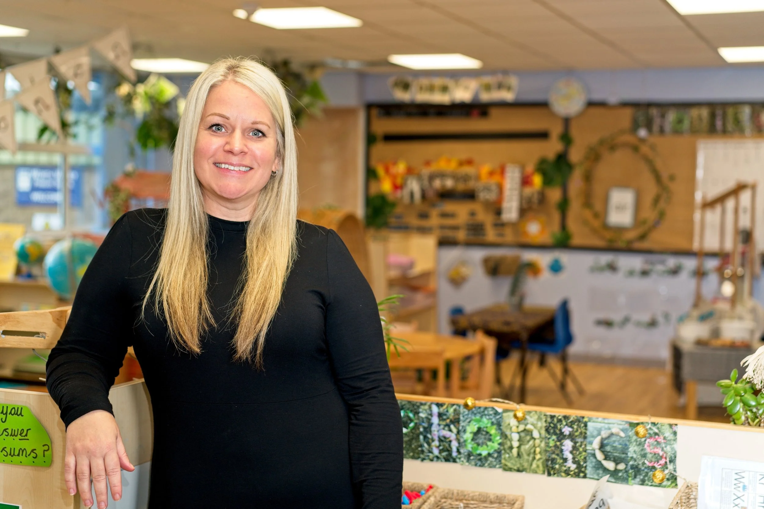 A smiling woman with long blonde hair wearing a black long-sleeve shirt standing in a colourful indoor craft store or classroom with shelves, decorations, and furniture in the background. School photography by Southam based photographer.