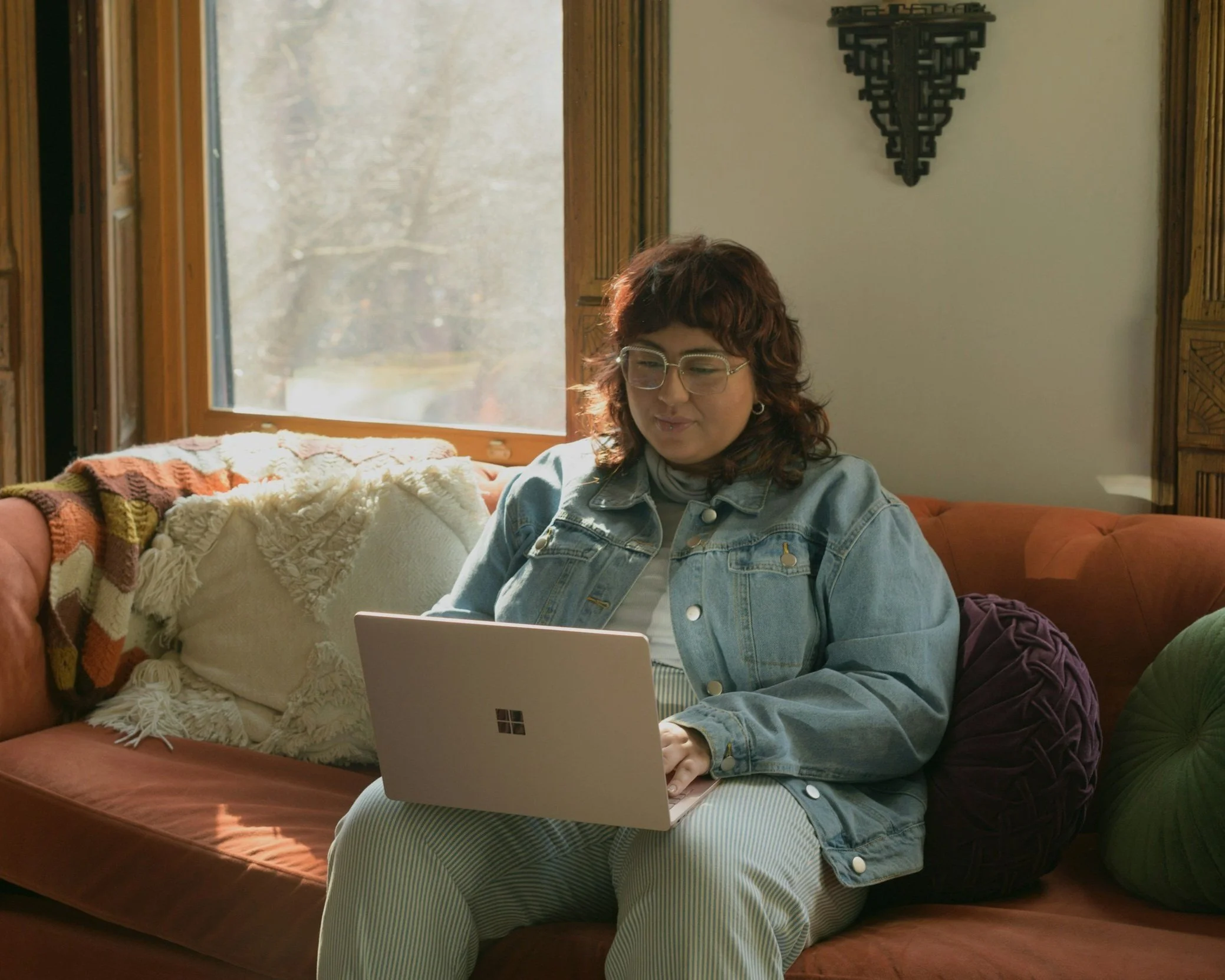 A person with long red hair with a jean jacket on and striped pants sitting on an orange couch working on their computer.