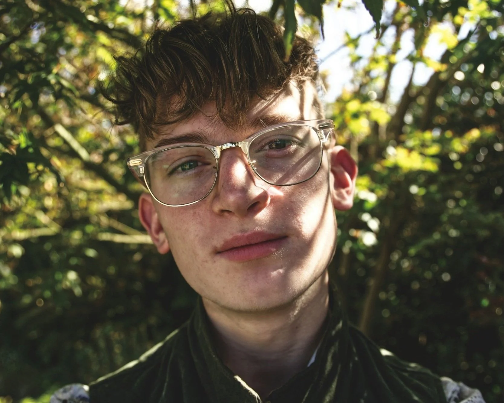 Gender non-confirming person with short brown-blonde hair wearing clear glasses with gold trim wearing a green jacket looking pleasantly curious at the camera