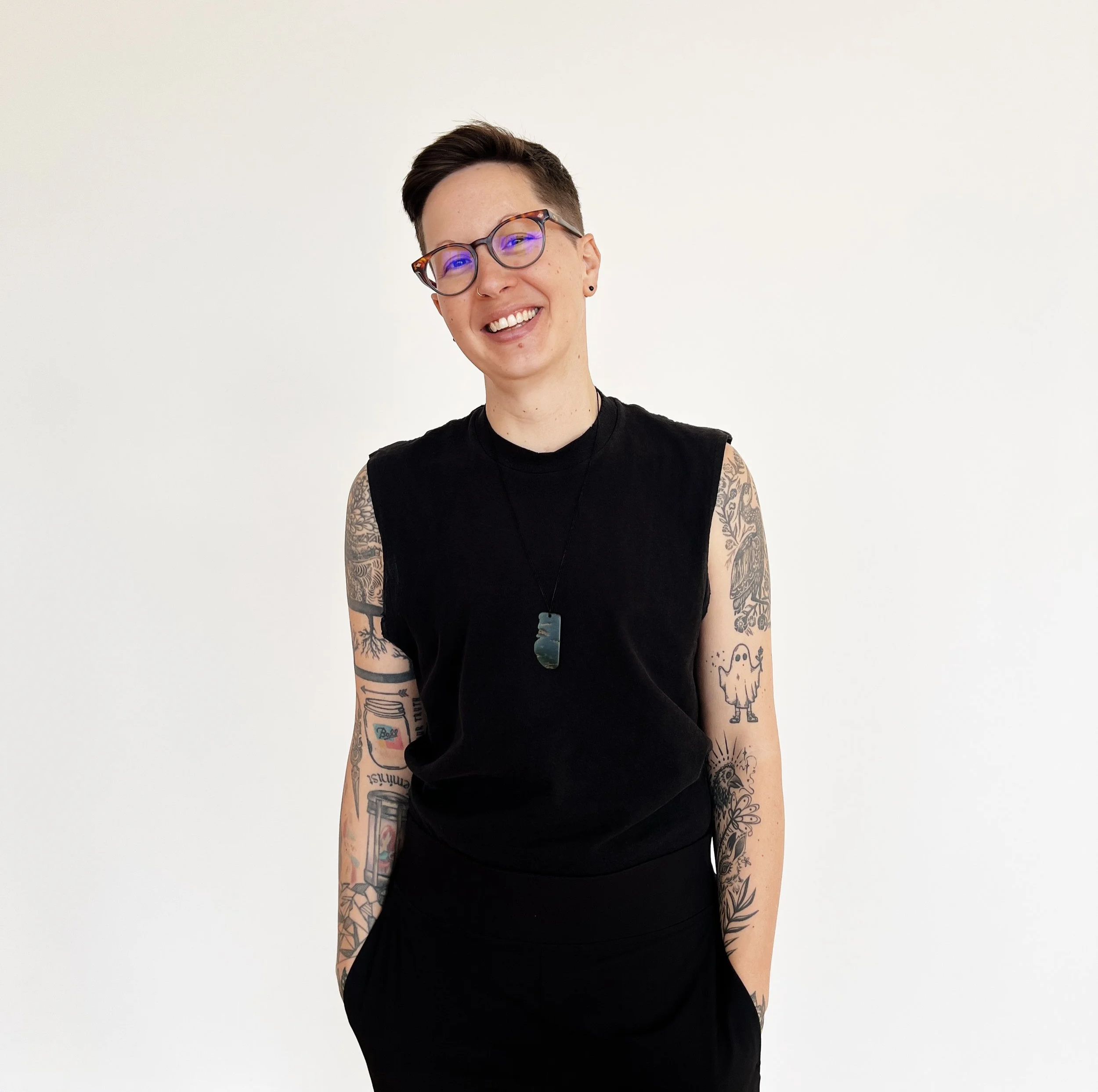 A smiling person with short dark hair, glasses, and tattoos on both arms, wearing a sleeveless black shirt and necklace, standing against a plain white background.