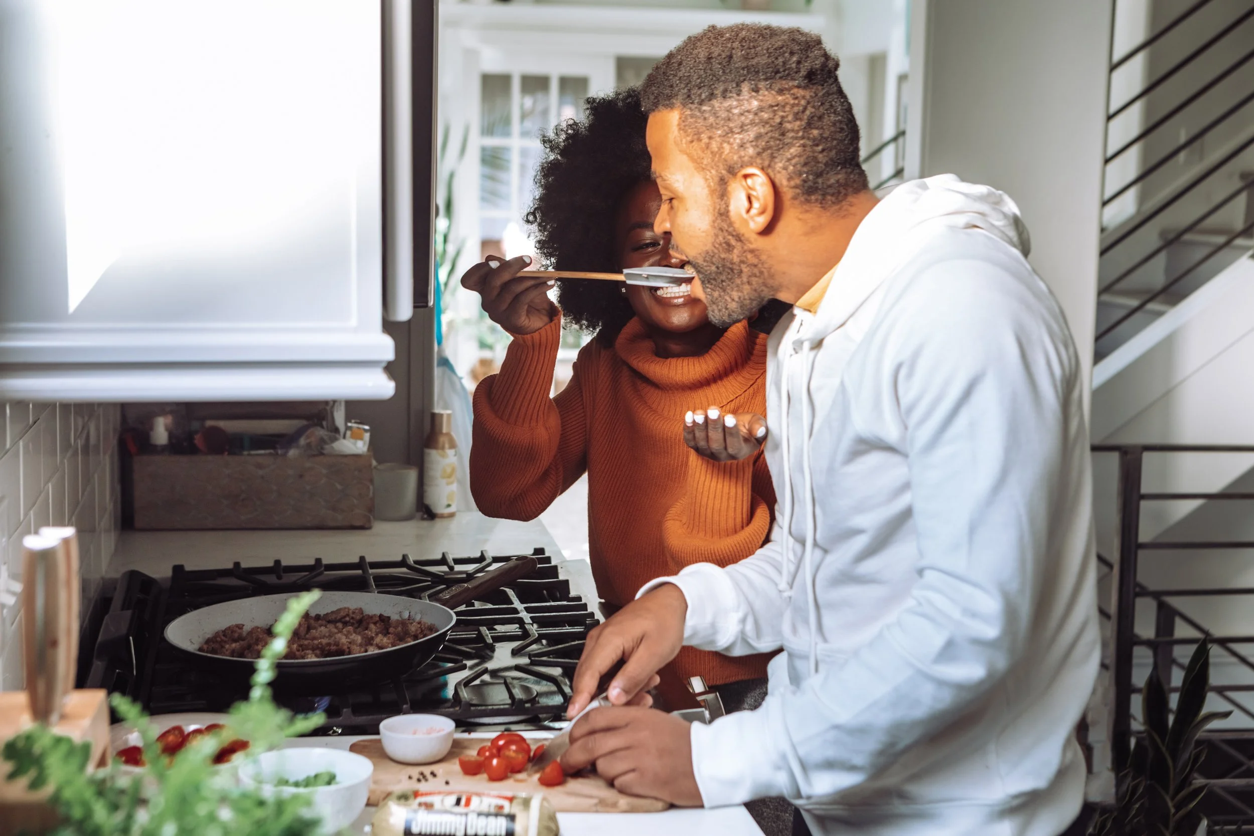 A man and woman cooking together in a kitchen, with the woman playfully feeding the man with a spoon.