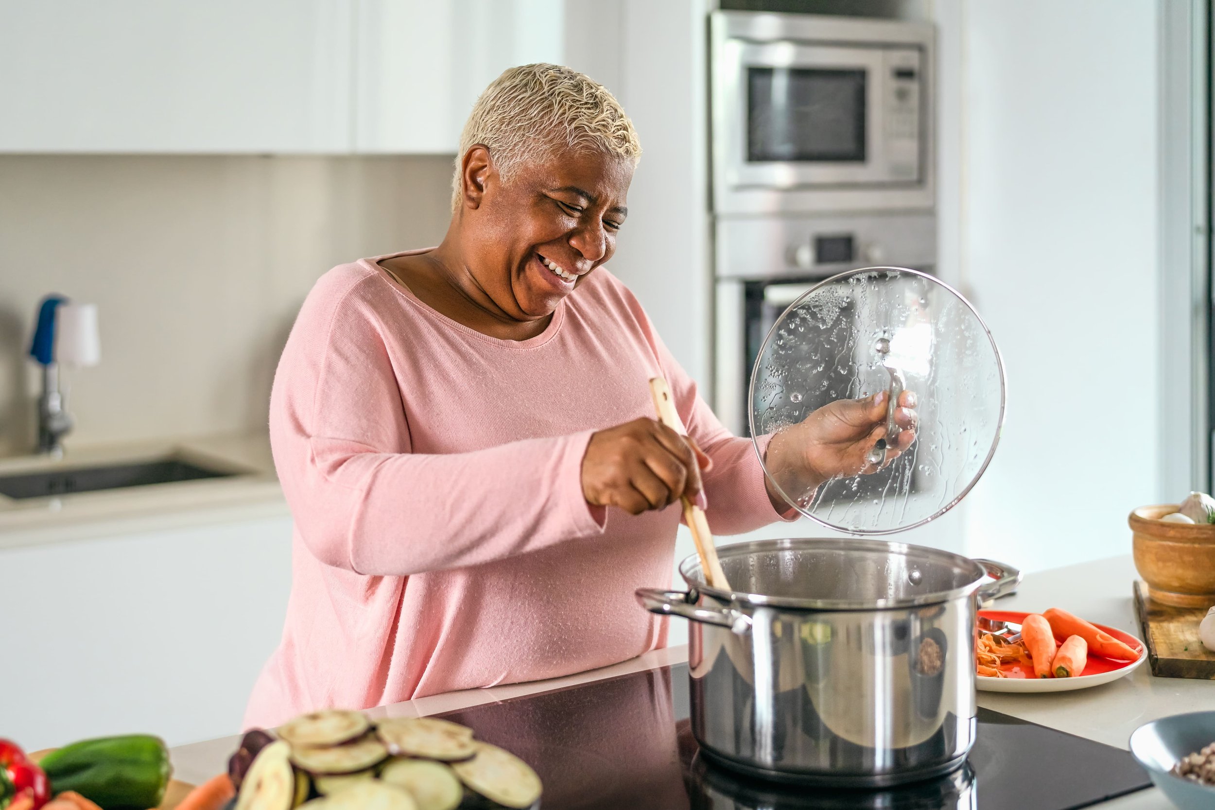 A woman smiling and cooking on a stovetop in a modern kitchen, holding a glass lid and stirring a pot with a wooden spoon. There are fresh vegetables like eggplants, carrots, and peppers on the counter.