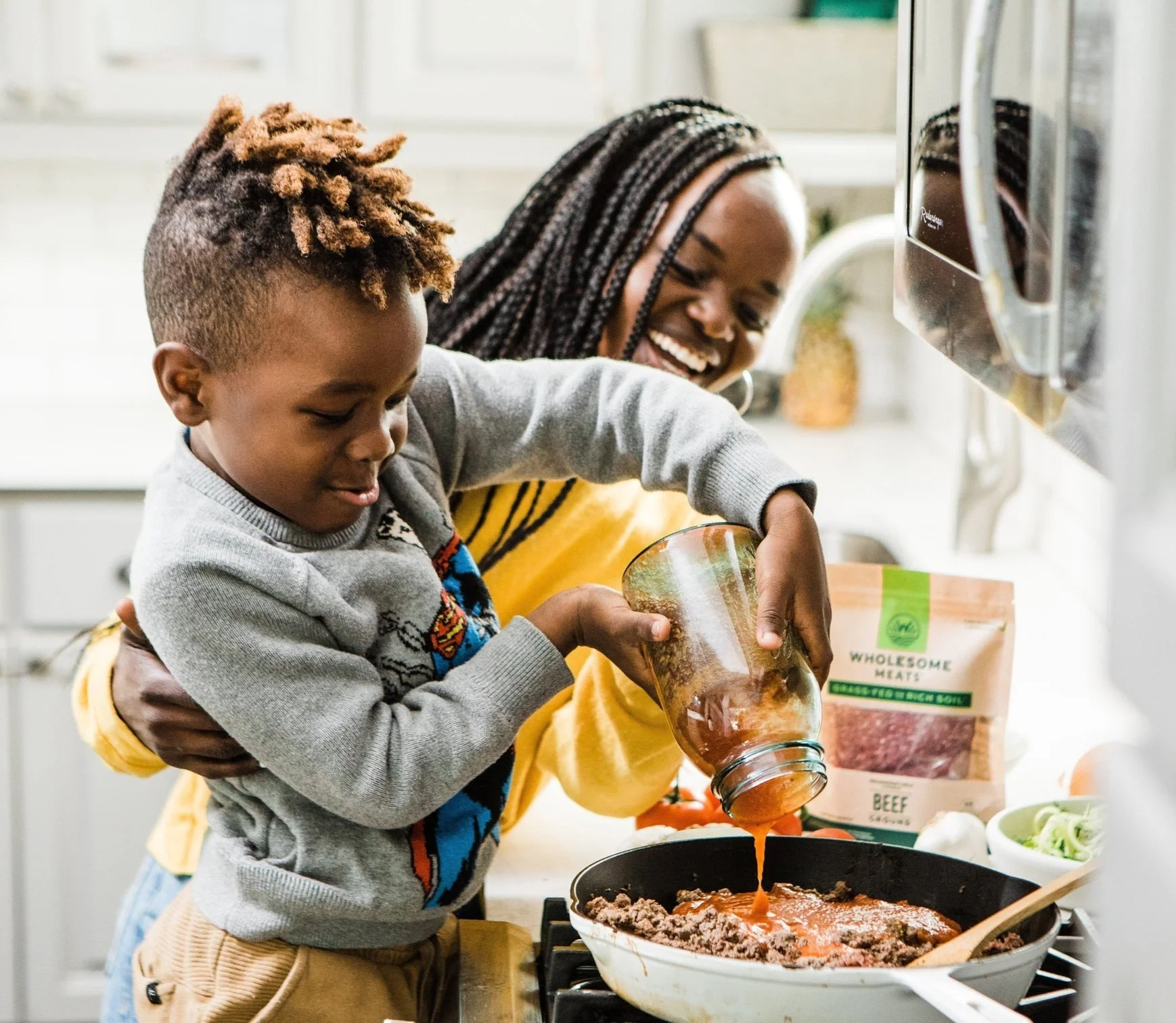 A woman and a young boy cooking together in a kitchen. The woman is smiling as she watches the boy pour tomato sauce into a frying pan.