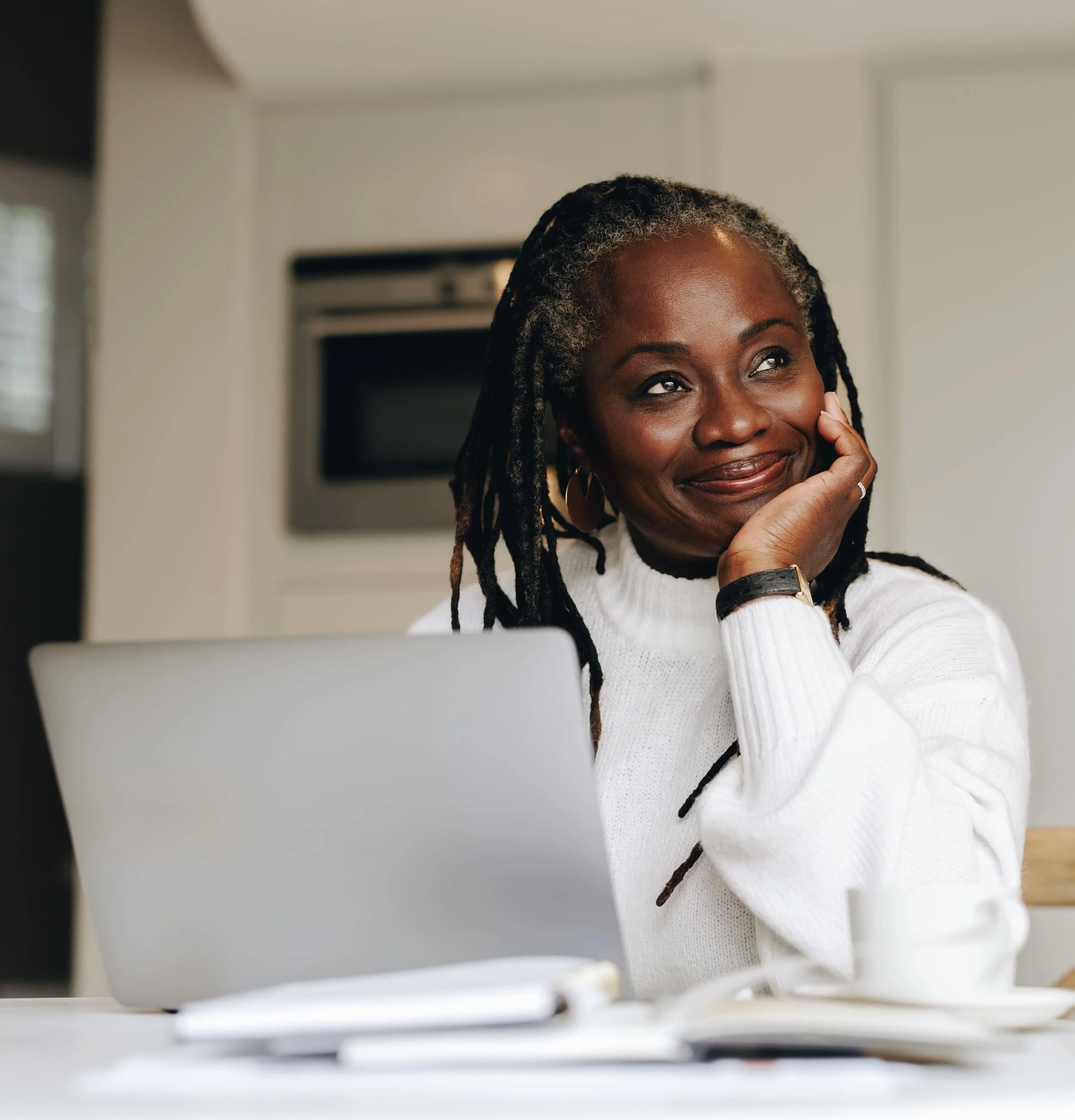 A smiling woman with dreadlocks wearing a white sweater, sitting at a desk with a laptop in front of her, in a modern kitchen.