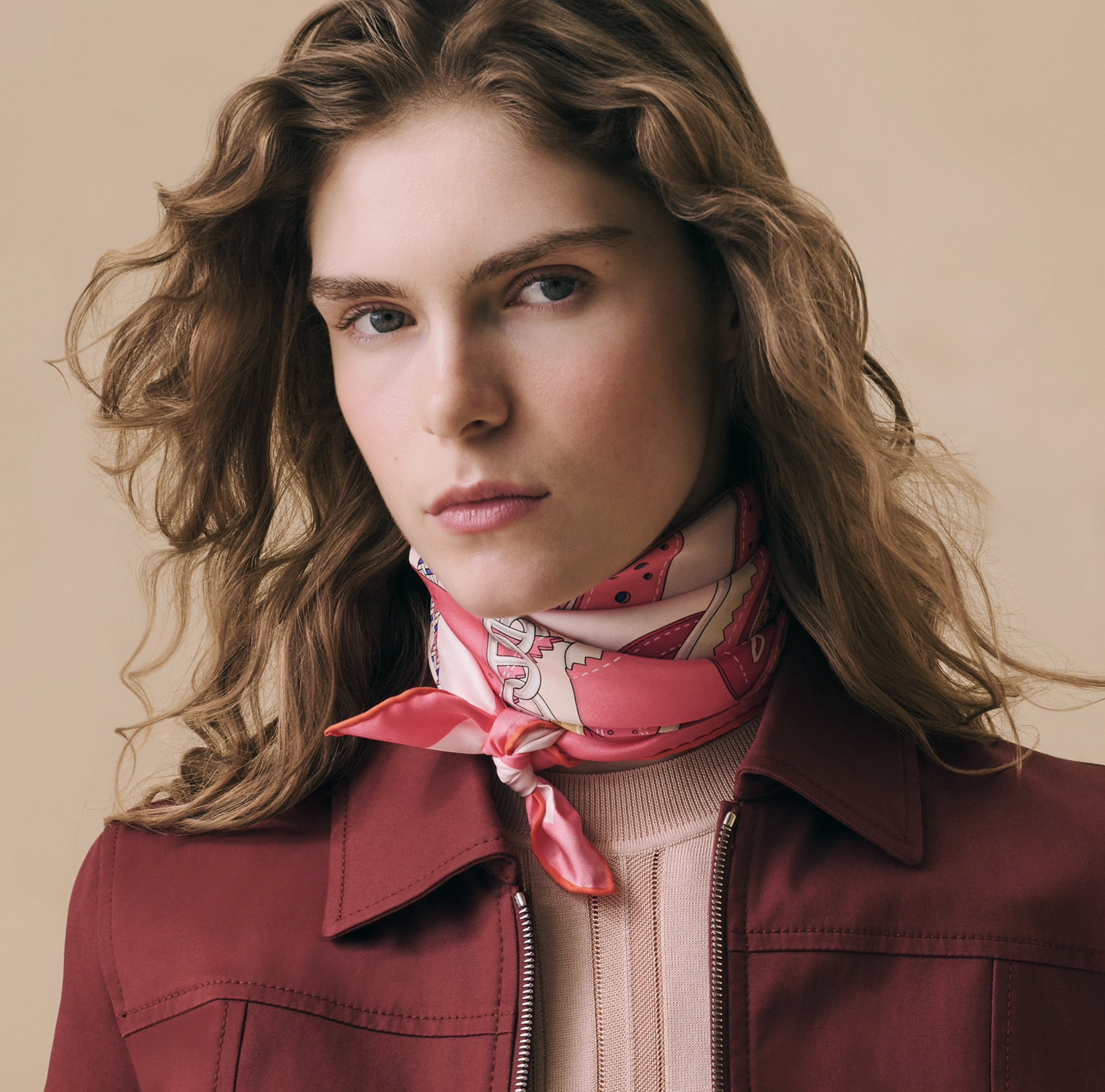 Cette photographie de mode en gros plan présente une mannequin aux longs cheveux châtains clairs, ondulés et volumineux, posant de face devant un arrière-plan neutre de couleur beige sablonneux. Elle regarde l'objectif avec une expression sereine et 