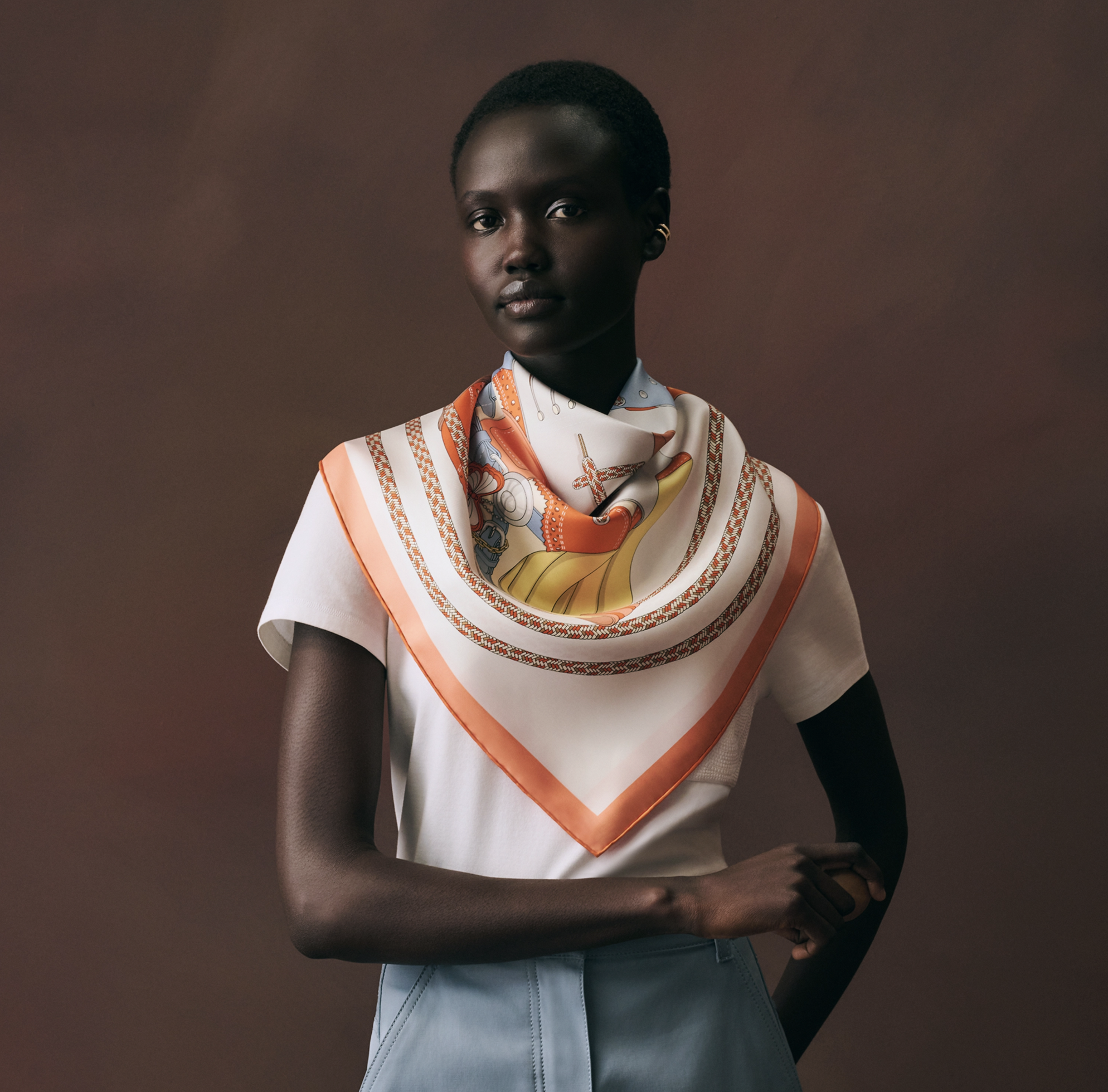 Portrait d'un mannequin femme noire à la coupe afro courte, posant devant un fond brun chaud et terreux. Elle porte le carré de soie 'derby_2000_foulard_hermes_4.png' noué élégamment autour du cou. Le foulard est majoritairement blanc cassé avec une 