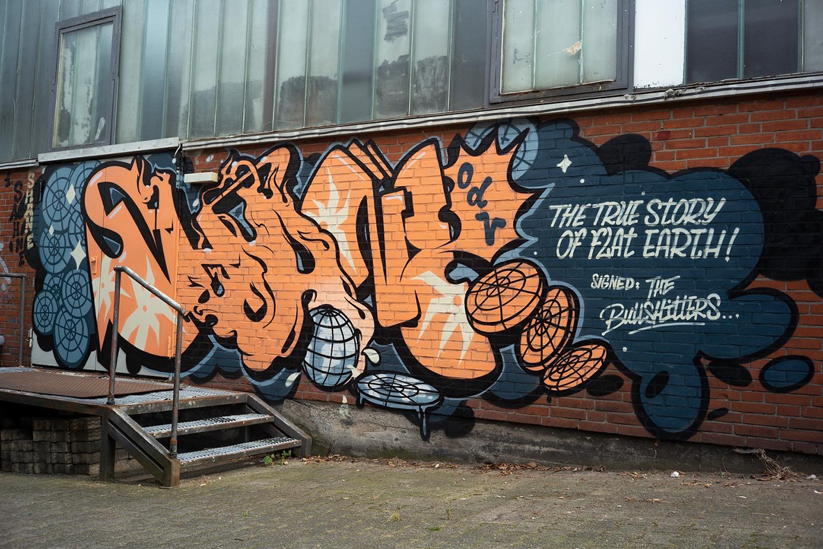 Fresque graffiti de l'artiste Shane Hello peinte sur un mur de briques rouges au pied d'un bâtiment industriel. Le lettrage orange vif « SHANE » repose sur un fond sombre découpé avec une terre plate, une sphère géométrique et la phrase écrite « THE 
