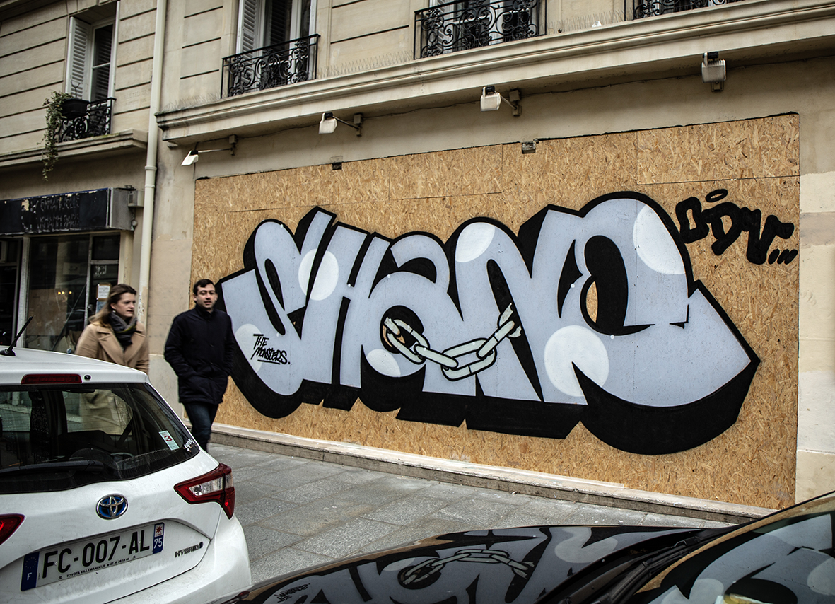Graffiti de lettrage argenté et noir par le graffeur Shane Hello sur un panneau de bois d'une façade parisienne. L'œuvre représente le nom « SHANE » écrit en pièces arrondies avec des bulles blanches, une chaîne en métal reliant deux lettres, et les 