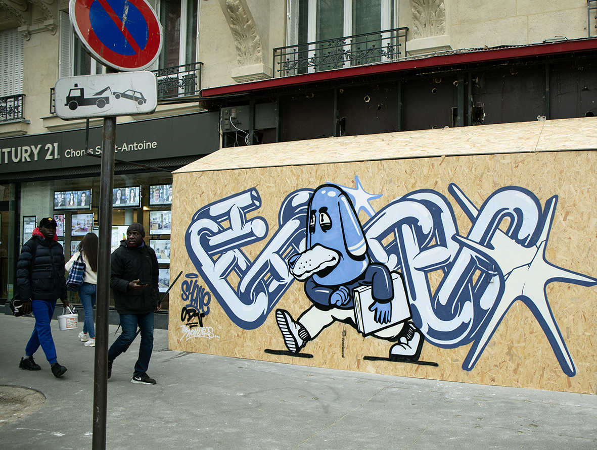 Street art sur un panneau de chantier en bois dans une rue parisienne par Shane Hello. L'œuvre en bleu et blanc montre un personnage de chien anthropomorphe marchant avec un livre sous le bras, intégré au milieu de maillons de chaînes entrelacés et d