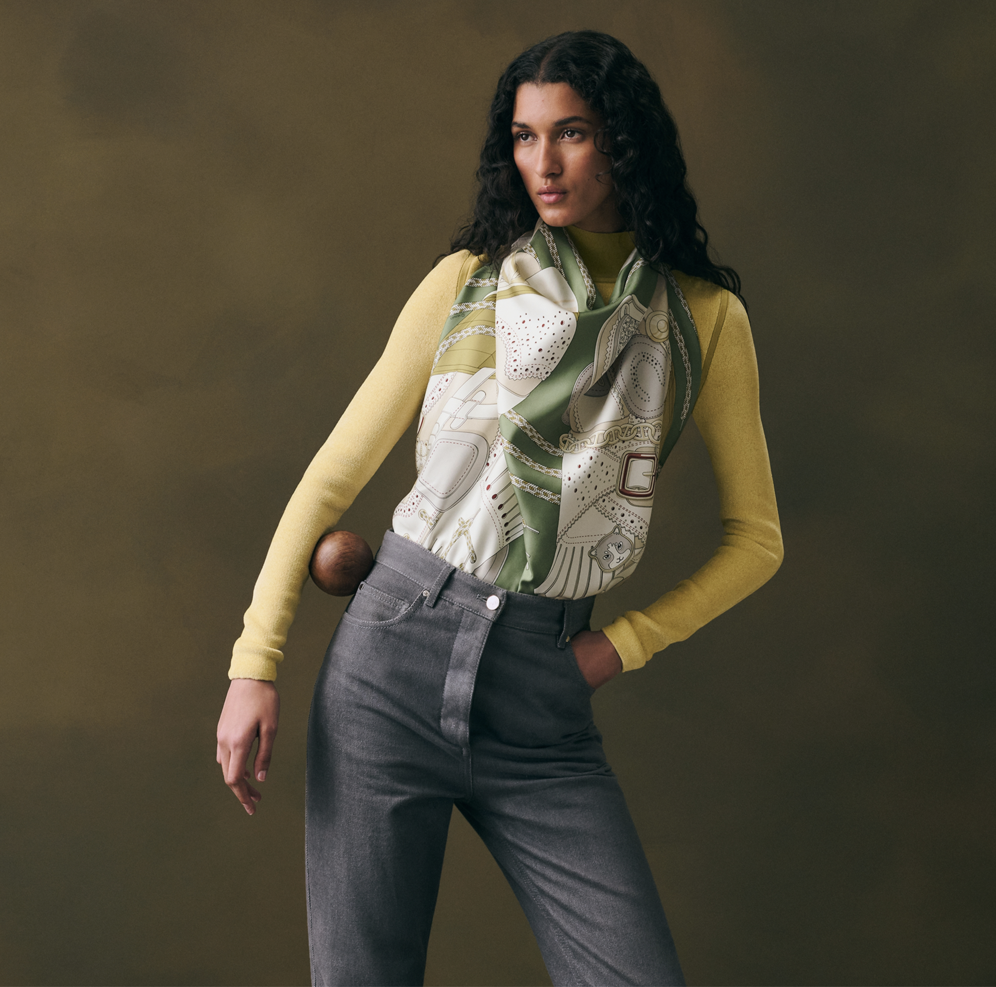 Cette photographie de mode en plan américain présente une mannequin aux longs cheveux bruns bouclés, posant de trois-quarts devant un arrière-plan texturé de couleur vert olive sombre. Elle porte un pull à col montant en maille fine jaune moutarde et