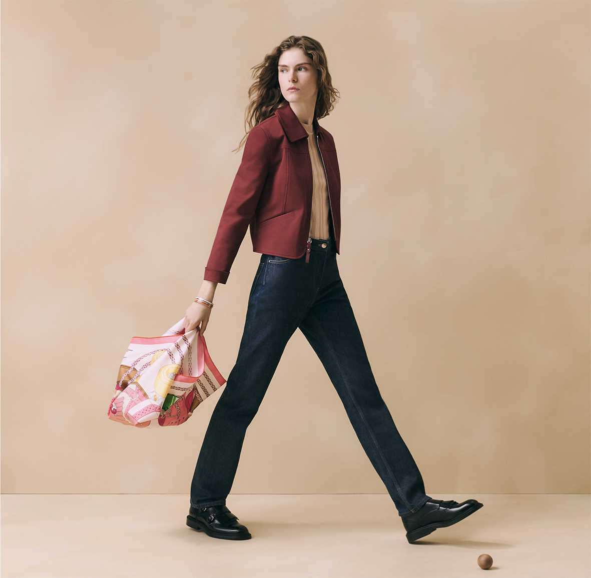 Modèle féminin marchand en studio dans une tenue élégante (veste bordeaux, jean brut), tenant à la main le carré en soie Hermès "Derby 2000" rose et jaune illustré par Shane hello.