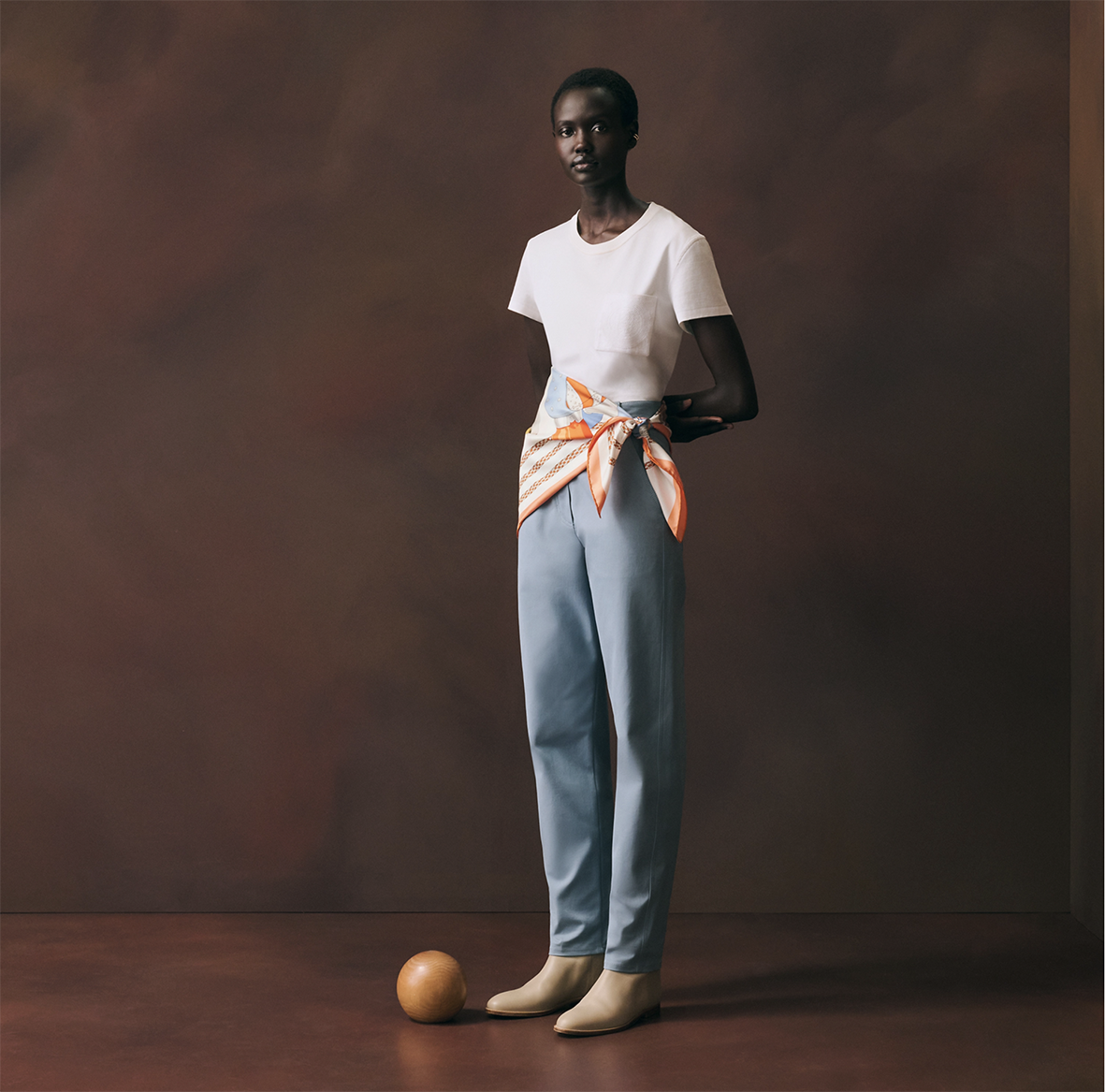 Modèle féminin posant en pied dans un studio brun, portant le carré en soie "Derby 2000" de Shane hello noué à la taille comme une ceinture, avec un t-shirt blanc et un pantalon bleu ciel.