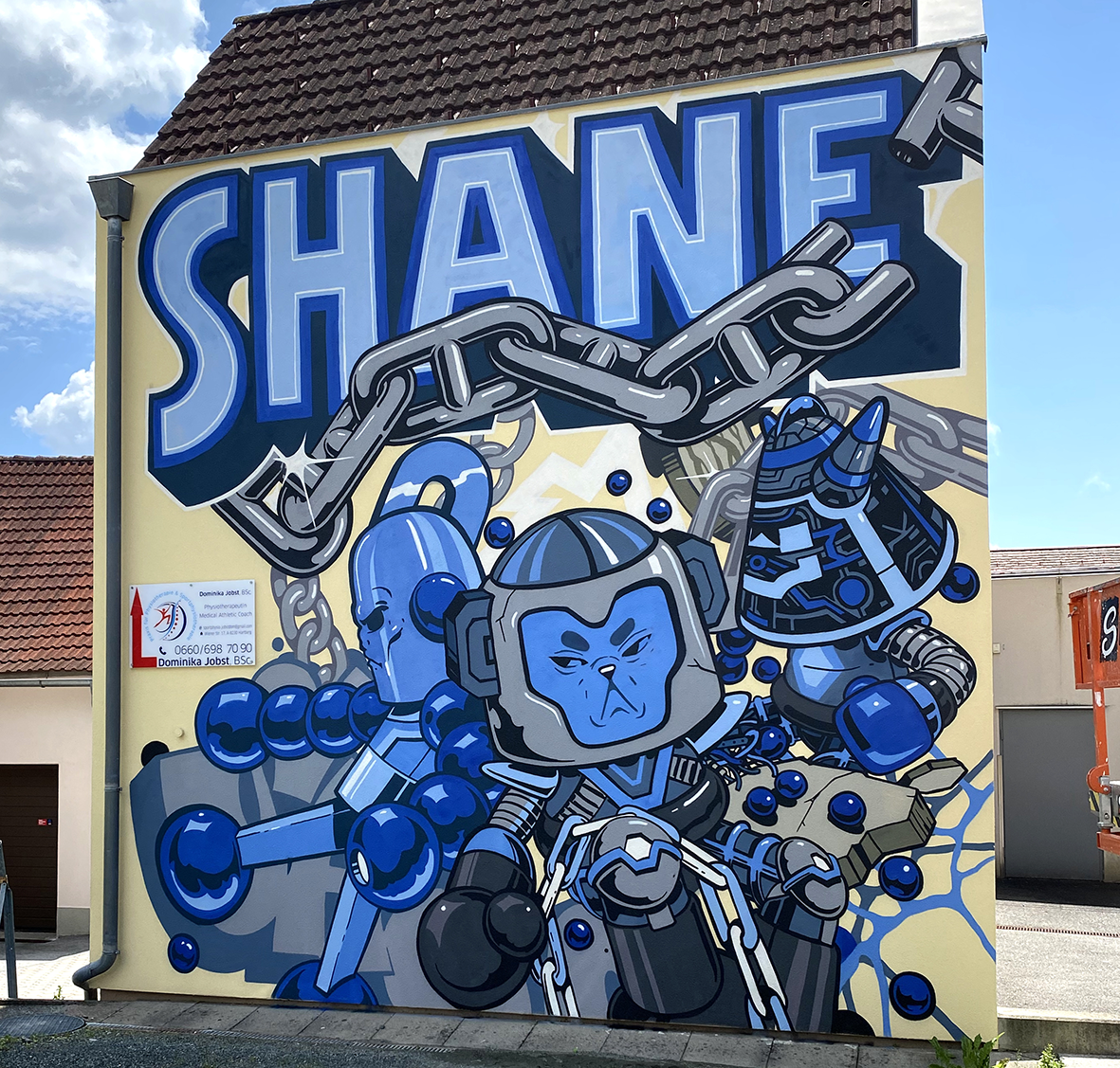 Fresque murale de street art par Shane Hello sur la façade d'un bâtiment. L'œuvre en camaïeu de bleus, gris et noir sur fond jaune présente le lettrage géant « SHANE » au sommet, traversé par une chaîne métallique grise. Au-dessous se trouvent trois 
