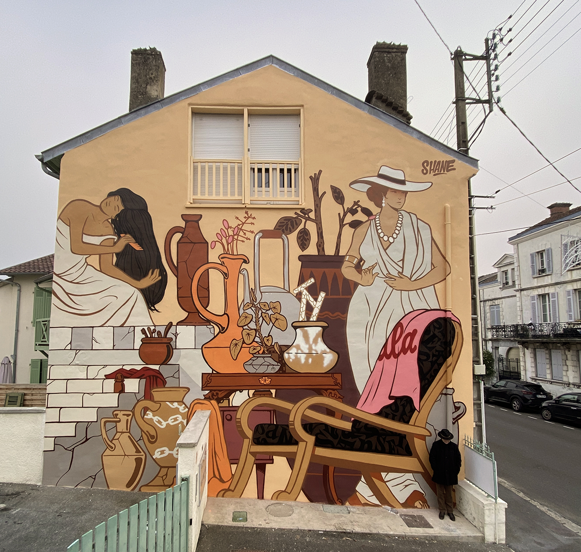 Fresque murale monumentale de street art peinte par Shane Hello sur la façade pignon d'une maison. L'œuvre aux teintes ocre, terre cuite, marron et blanc crème illustre une scène d'inspiration antique et méditerranéenne : une femme se peignant les ch