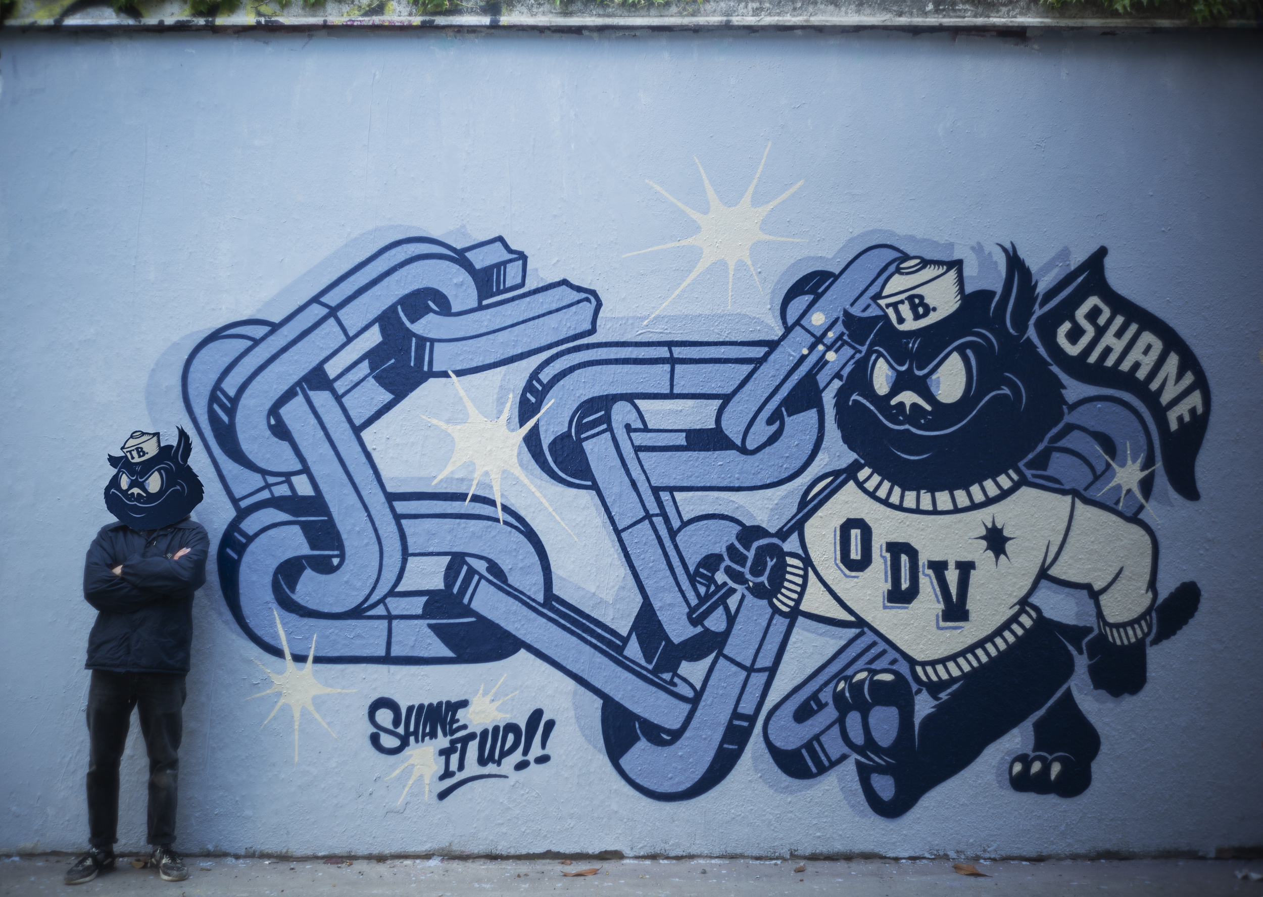 Fresque murale de street art réalisée par Shane Hello sur un mur bleu. L'œuvre présente d'imposantes chaînes métalliques stylisées en 3D dans un camaïeu de bleus, accompagnées d'un personnage de chat noir cartoon habillé d'un pull crème marqué « ODV 