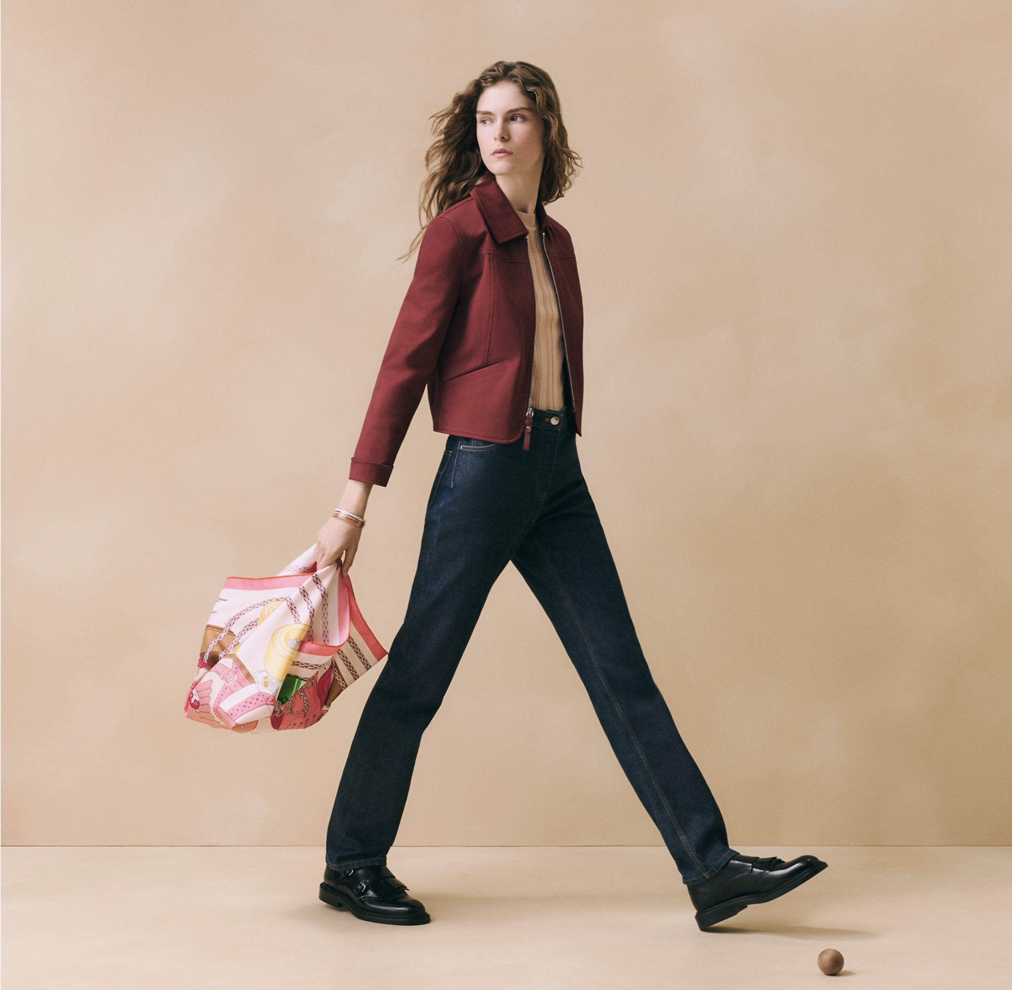 Cette photographie de mode en pied montre une mannequin en mouvement, marchant de profil vers la droite avec une enjambée dynamique. Elle est mise en scène devant un arrière-plan studio épuré de couleur beige ocre doux. La mannequin, aux longs cheveu