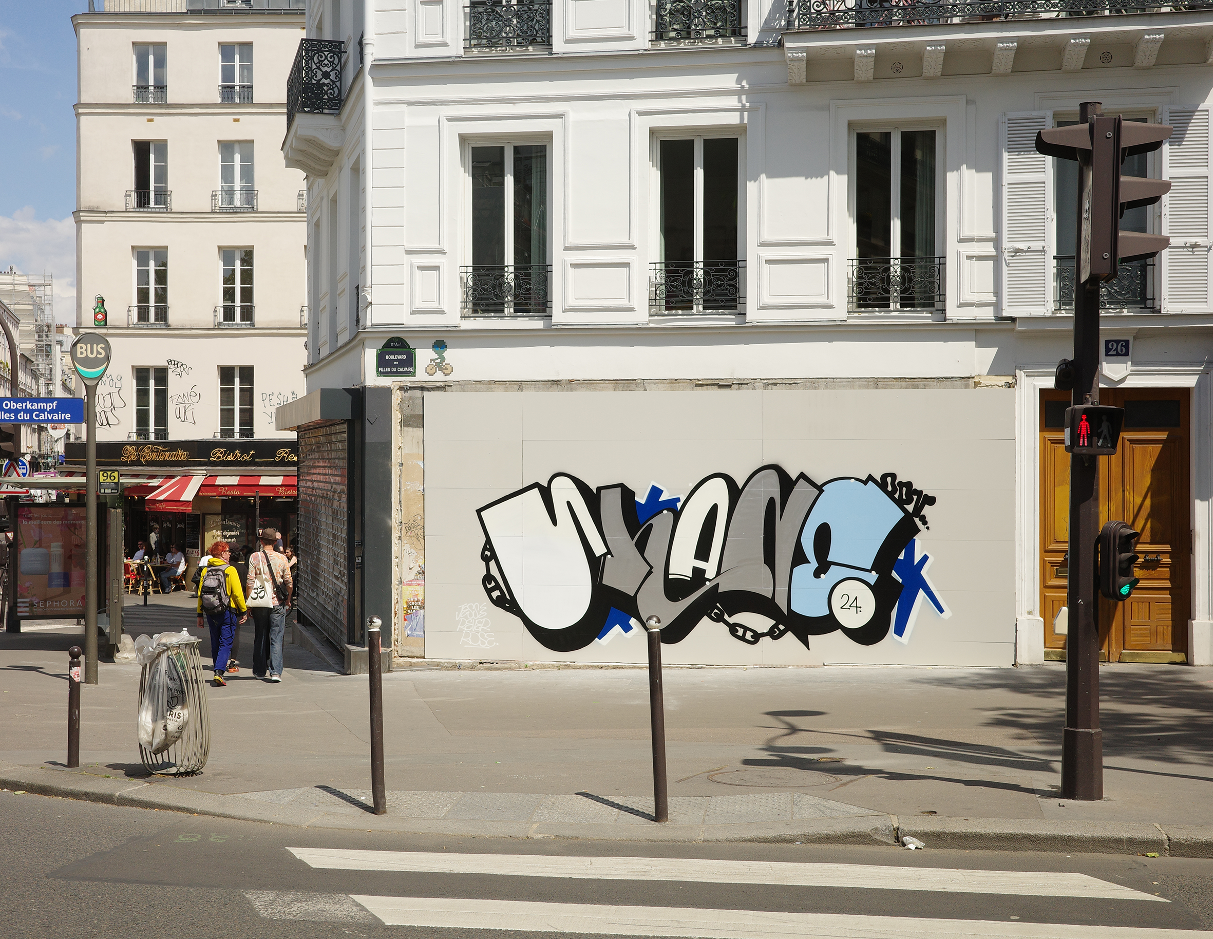 Graffiti de street art par Shane Hello sur un panneau blanc d'un immeuble haussmannien à l'angle du Boulevard des Filles du Calvaire à Paris. L'illustration représente le lettrage « SHANE » en blanc, gris et bleu ciel avec des motifs de chaînes métal