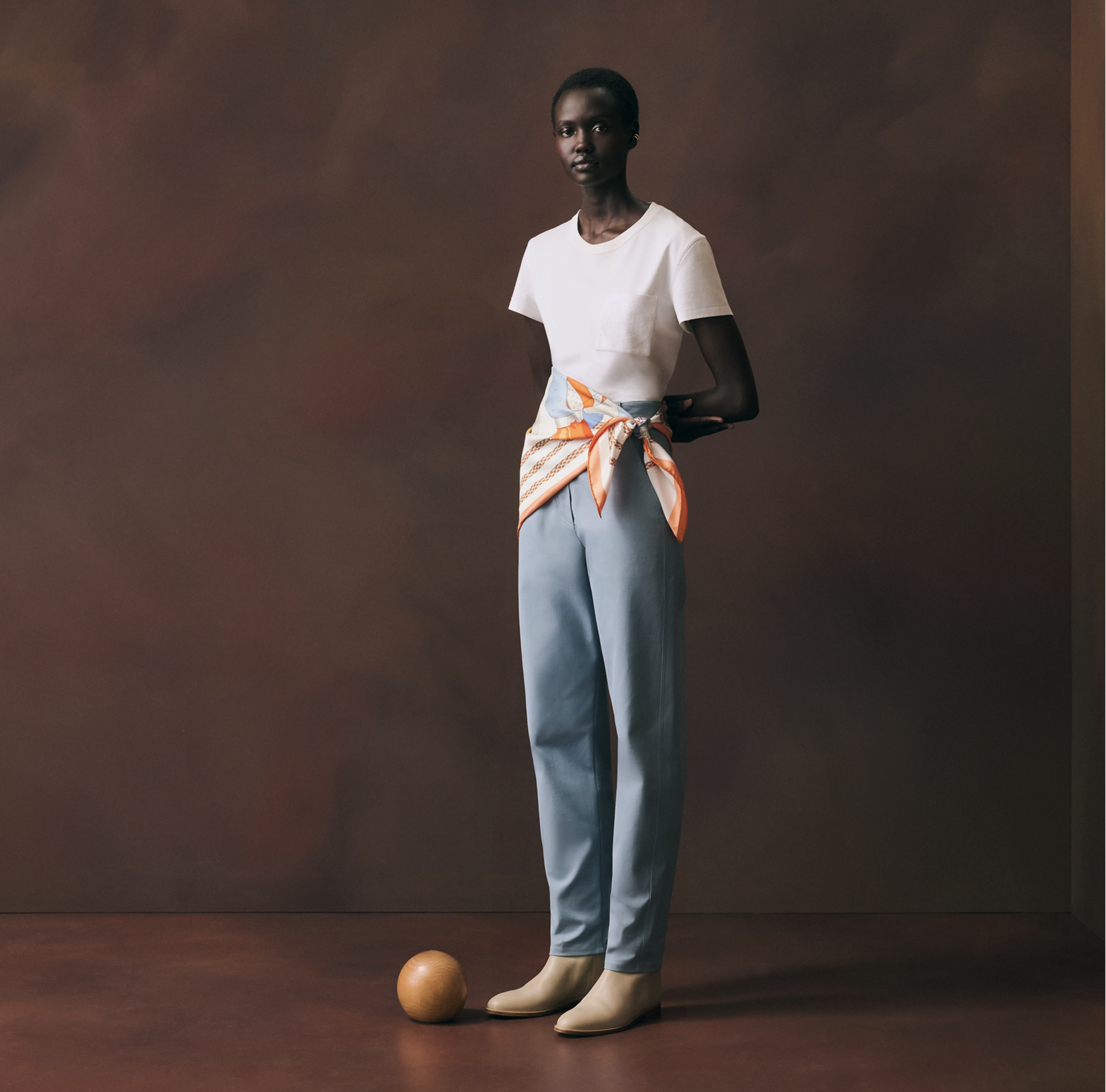 Cette photographie en pied présente un mannequin femme noir au port élégant, debout et de face, contre un arrière-plan d'atelier texturé d'un brun riche et terreux. Il porte une tenue minimaliste composée d'un t-shirt blanc uni et d'un pantalon taill