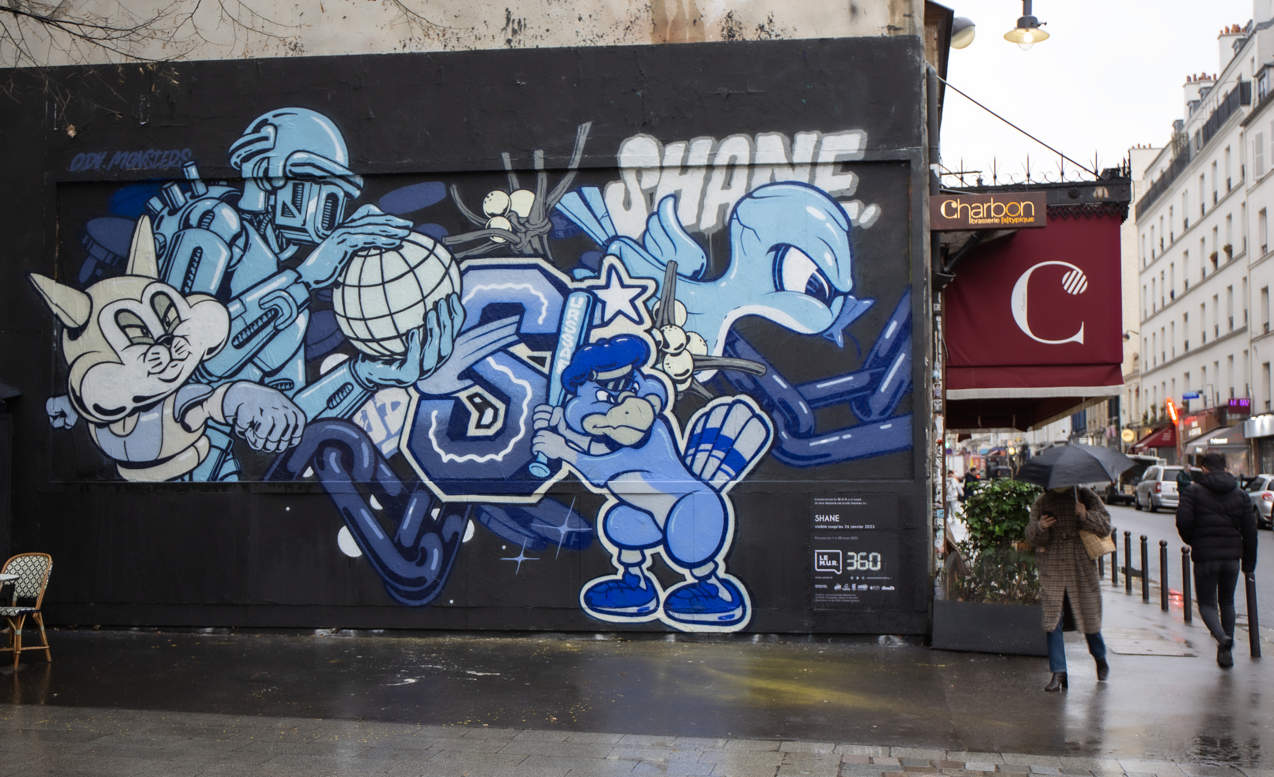 Fresque street art en camaïeu de bleus réalisée par Shane Hello sur Le M.U.R Oberkampf (n° 360) à Paris, situé à côté du café Charbon. L'œuvre sur fond noir rassemble plusieurs personnages cartoon : un chat stylisé, un personnage en combinaison spati
