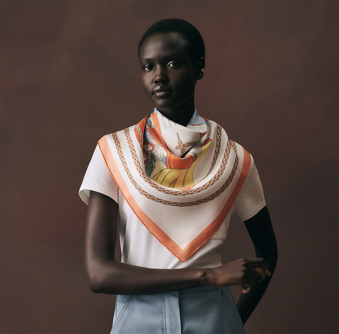 Modèle féminin portant le foulard en soie Hermès "Derby 2000" conçu par l'illustrateur Shane hello, noué élégamment sur un t-shirt blanc et un pantalon bleu, révélant la bordure orange et le motif graphique rétro.
