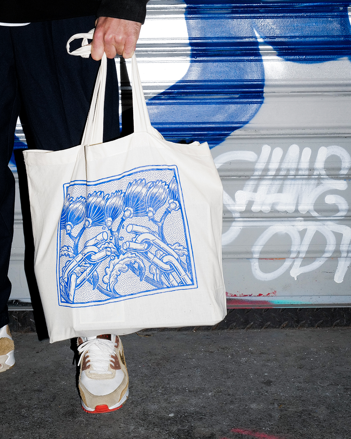 Sac tote bag en coton naturel beige porté à la main par une personne en tenue streetwear, devant un rideau de fer graffé. Le sac présente une illustration carrée sérigraphiée en bleu cobalt par l'artiste Shane Hello, représentant des fleurs stylisées
