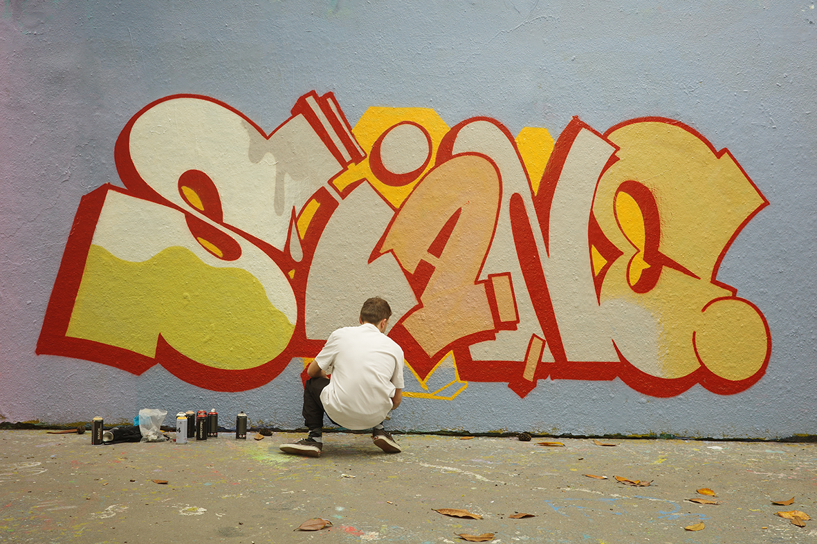 L'artiste graffeur Shane Hello de dos, accroupi en train de peindre une fresque graffiti sur un mur extérieur. Le lettrage volumineux « SHANE » affiche des teintes chaudes jaunes, beige et pêche, soulignées par un contour rouge vif sur fond bleu past