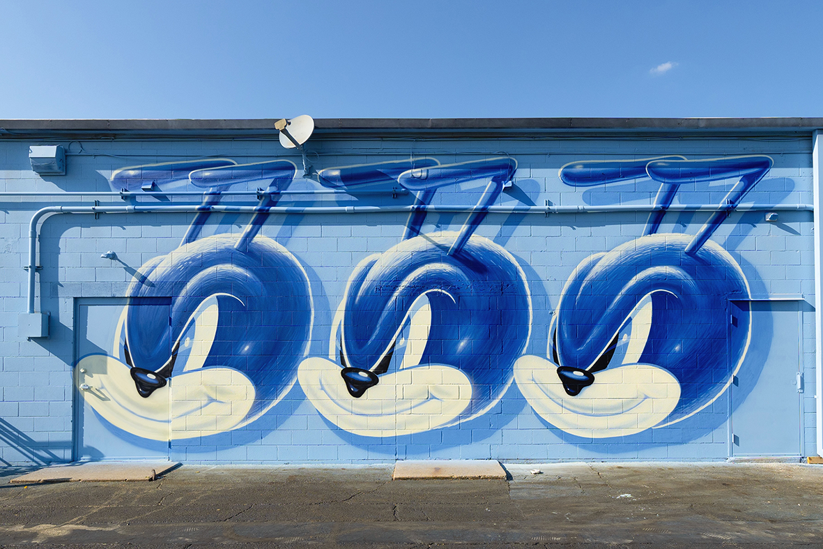 Fresque murale monumentale de street art par Shane Hello sur la façade bleue d'un bâtiment en parpaings. L'œuvre présente un motif répétitif composé de trois têtes identiques d'un personnage de moustique cartoon au regard malicieux, peint dans un cam