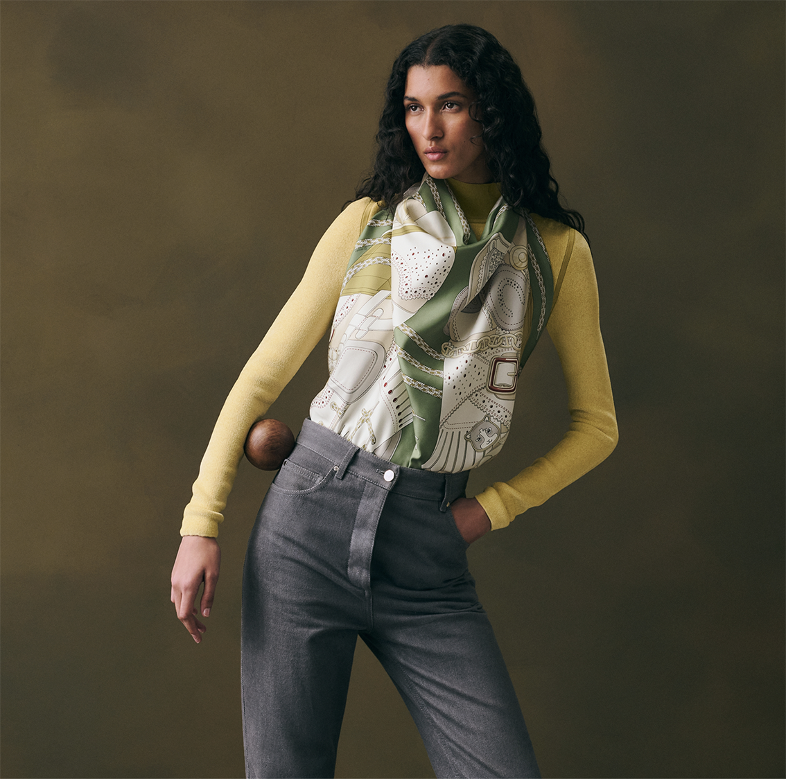 Modèle féminin posant en studio, portant le carré de soie Hermès "Derby 2000" vert sauge de Shane hello drapé en plastron sur un pull jaune moutarde et un jean gris foncé.
