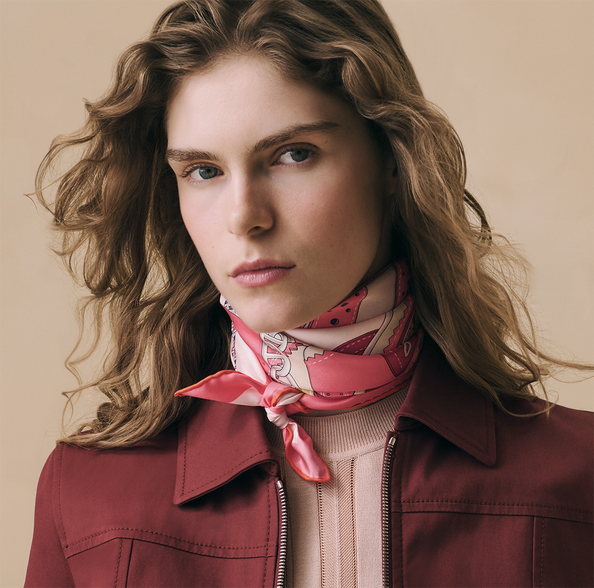 Portrait rapproché d'un mannequin féminin aux cheveux ondulés portant le carré en soie Hermès "Derby 2000" rose de Shane hello, délicatement noué autour du cou avec une veste bordeaux.