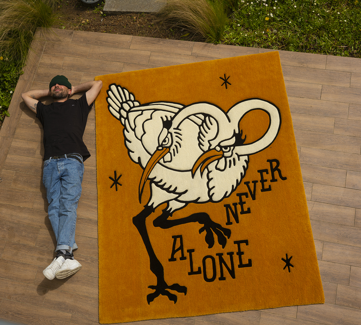 Vue en plongée d'un grand tapis tufté artisanal moutarde et crème créé par Shane Hello, posé sur une terrasse en bois à côté de l'artiste allongé. Le tapis illustre deux oiseaux à long bec (ibis ou hérons) aux cous entrelacés avec la typographie « NE