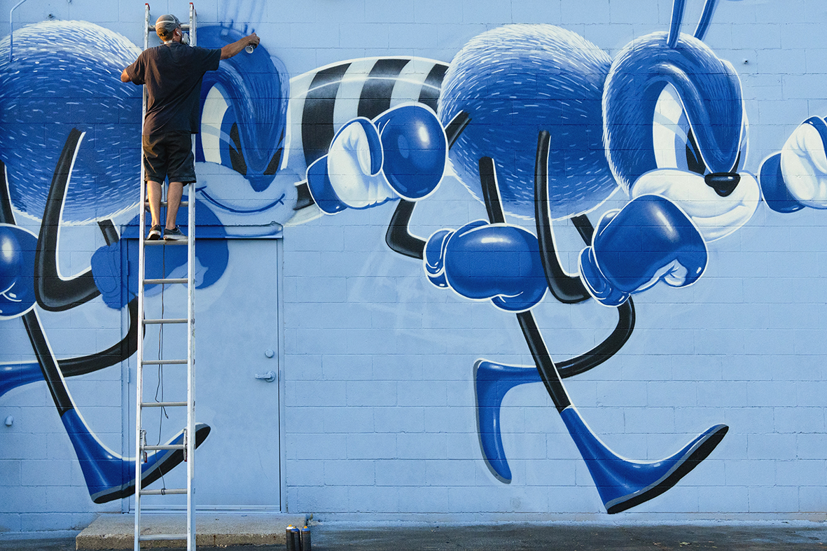 L'artiste graffeur Shane Hello perché sur une grande échelle en train de peindre à la bombe une fresque murale bleue sur un mur en parpaings. L'illustration représente des moustiques ou insectes cartoon géants en tenue de boxeur avec des gants bleus,
