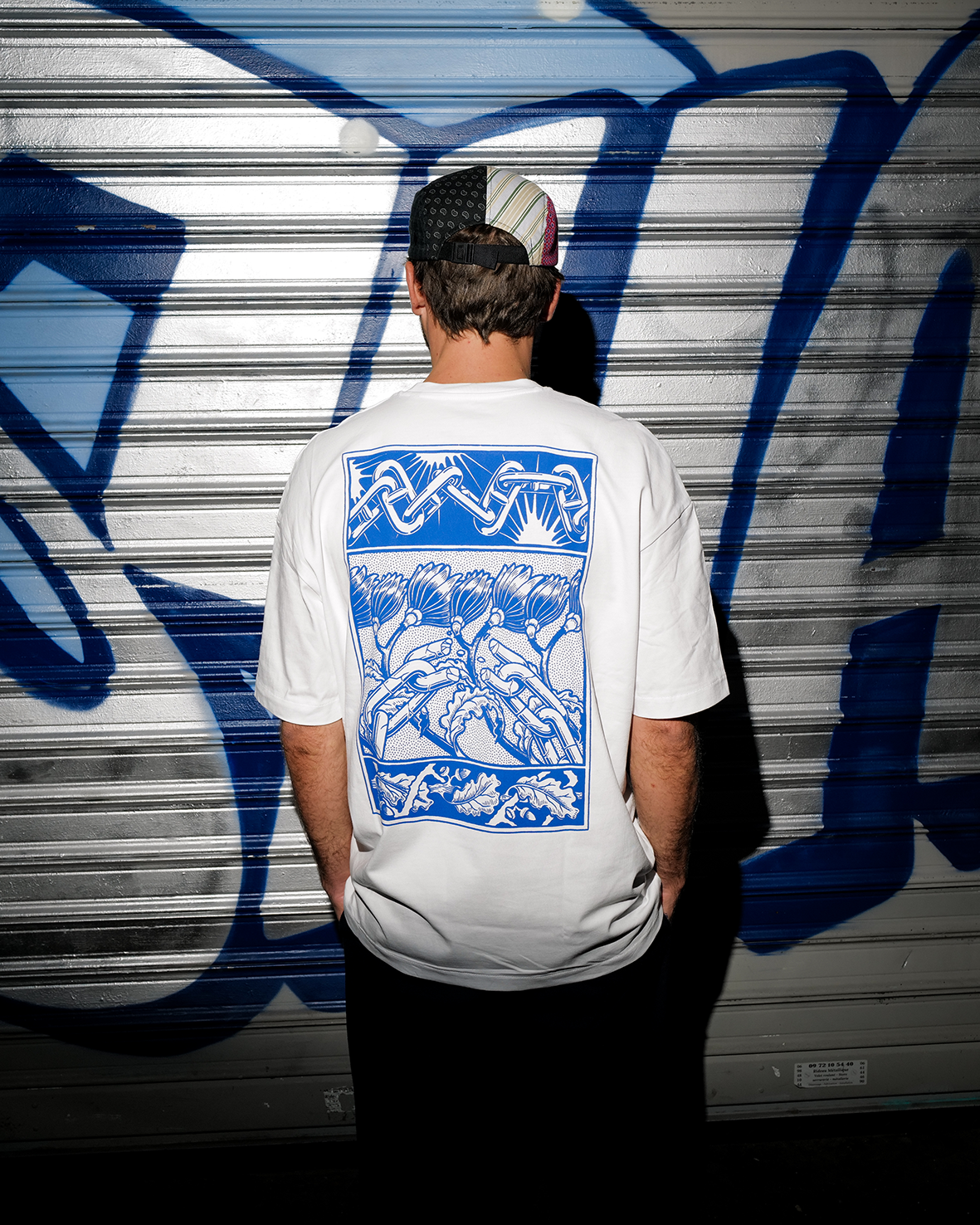 Vue de dos d'un homme portant un t-shirt oversize blanc de la marque Shane Hello, posant devant un rideau de fer graffé. Le dos du t-shirt présente une sérigraphie rectangulaire bleue imprimée d'une illustration d'inspiration Arts and Crafts : des ma