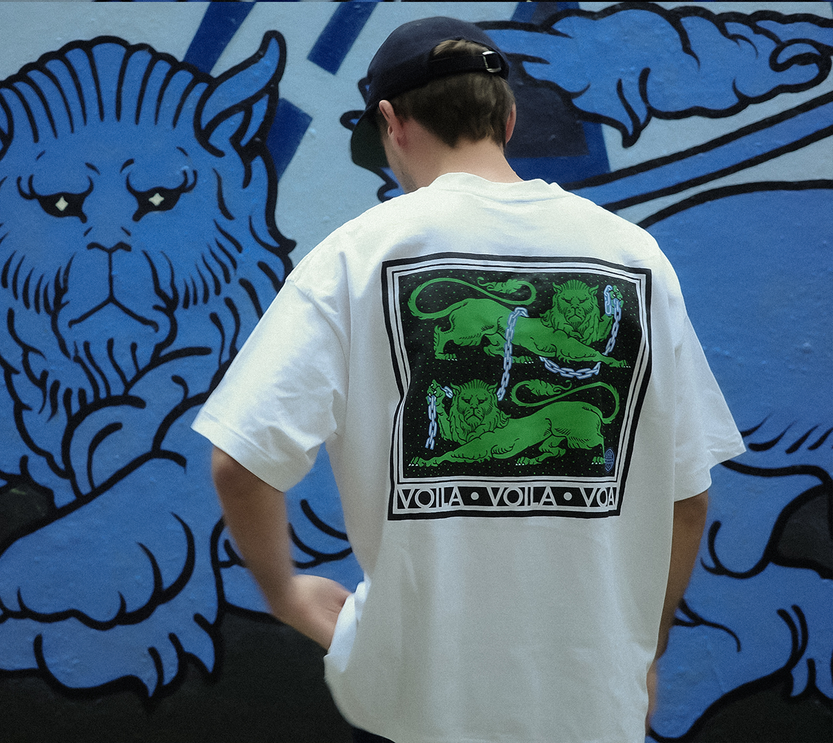 Gros plan de dos sur un homme portant le t-shirt blanc en coton de l'artiste Shane Hello devant une fresque murale bleue. L'imprimé au dos présente un carré noir encadré de blanc avec deux lions verts héraldiques reliés par des chaînes métalliques et