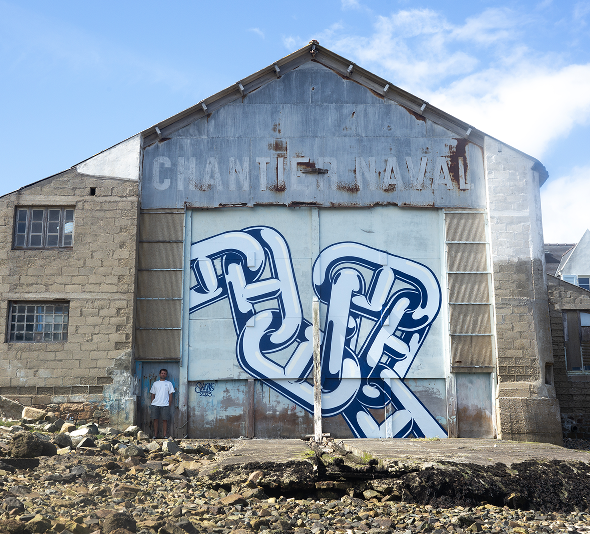 Fresque murale monumentale de l'artiste Shane Hello peinte sur la porte d'un hangar de chantier naval désaffecté en bord de mer. L'illustration représente un maillon de chaîne entrelacé géant bleu marine et blanc forming un lettrage stylisé. L'artist