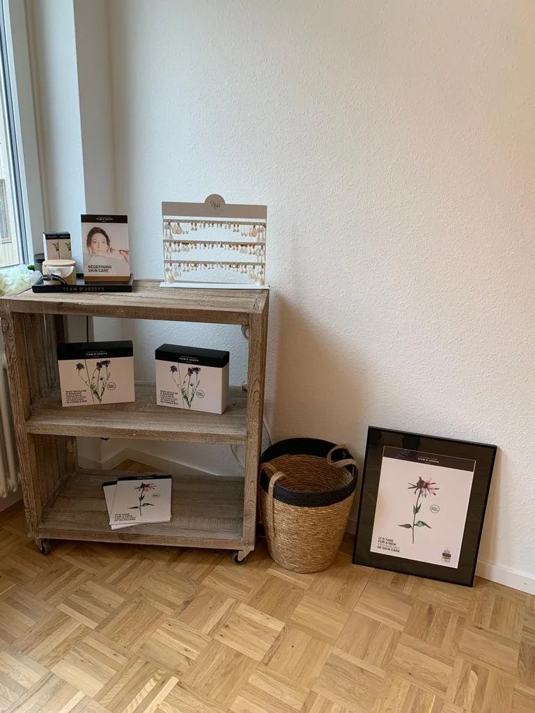Ein Holzregal mit Kosmetikprodukten und einer Displaykarte steht an einer weißen Wand. Neben dem Regal ist ein Korb und eine gerahmte botanische Illustration auf den Boden gelehnt. Die Produkte zeigen Bilder von Blumen im Zusammenhang mit Kosmetik.