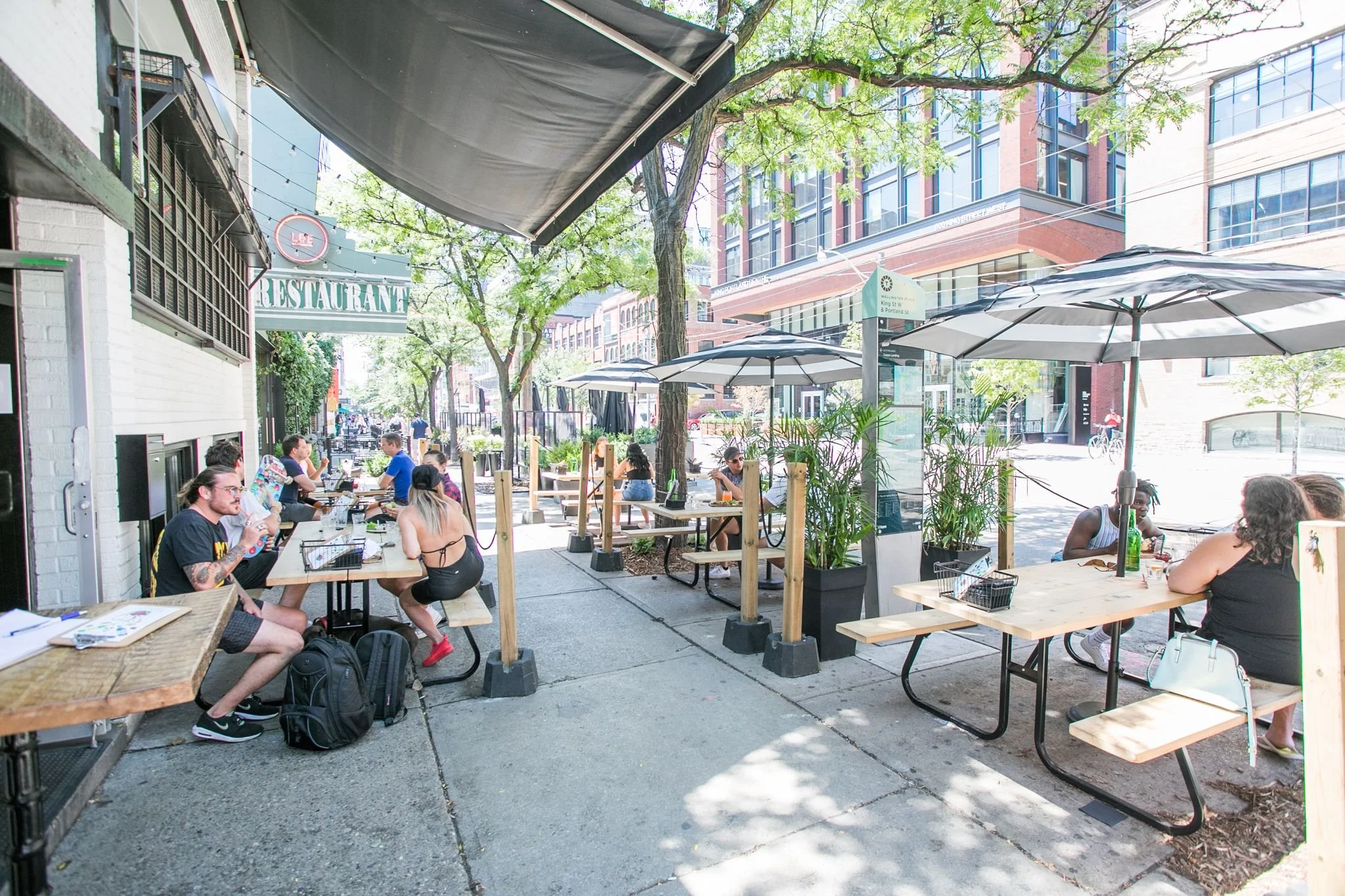 Outdoor patio of a restaurant with people sitting at picnic tables under large umbrellas, with a city street and tall buildings in the background.