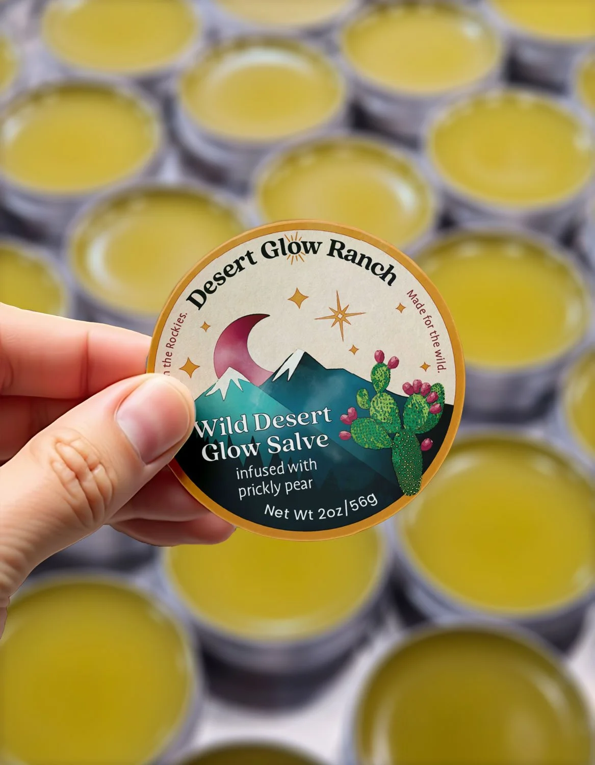 A hand holding a container of Wild Desert Glow Salve with a background of multiple yellow containers of the same product.
