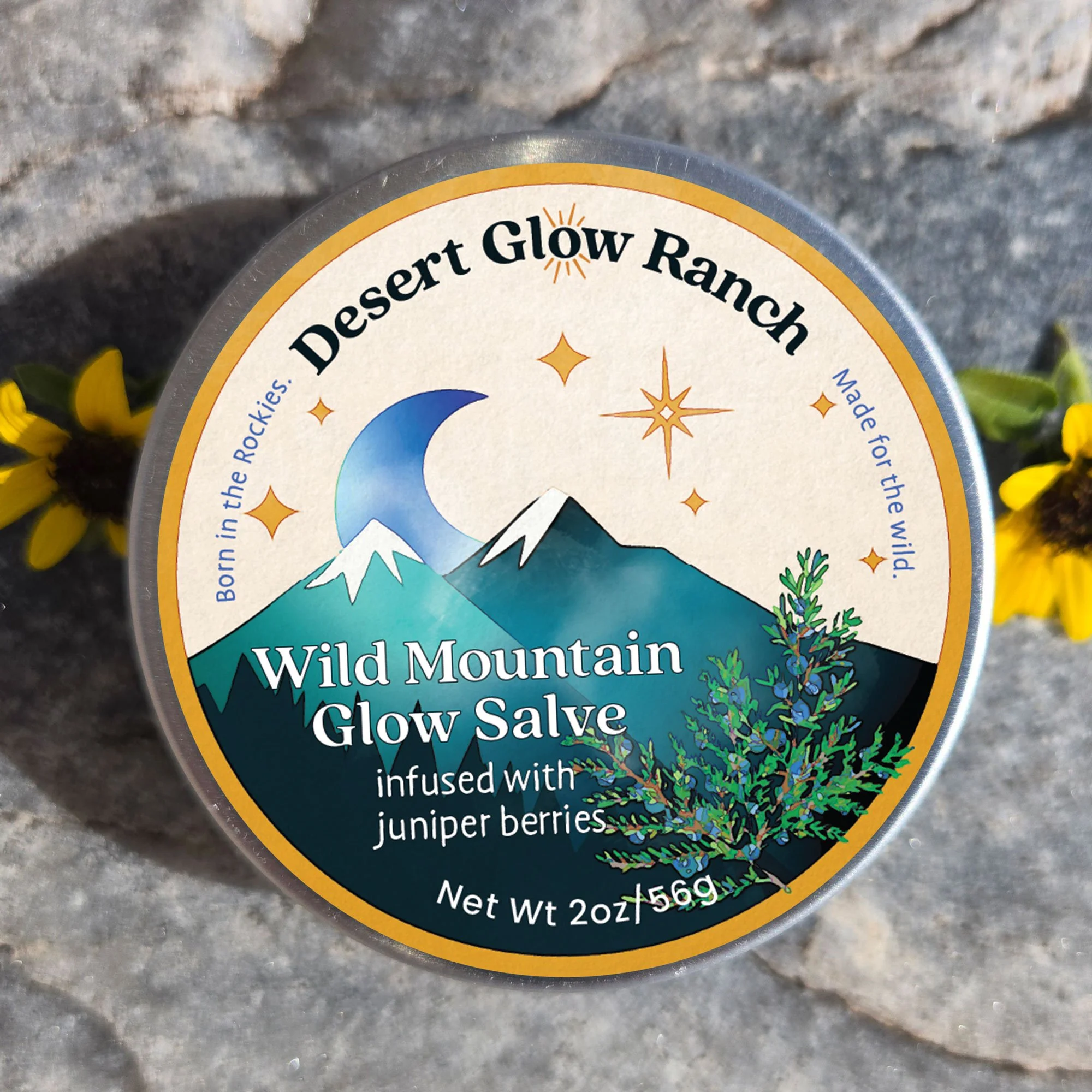 A container of Wild Mountain Glow Salve infused with juniper berries, with a mountain, moon, and star-themed label, placed on a rocky surface with yellow flowers nearby.