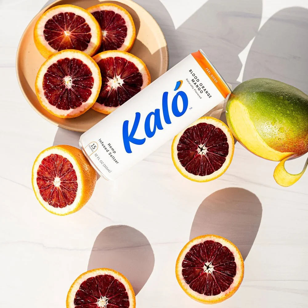 Blood orange and mango smoothie in a can labeled 'Kaló' with blood orange slices and a mango on a white surface.