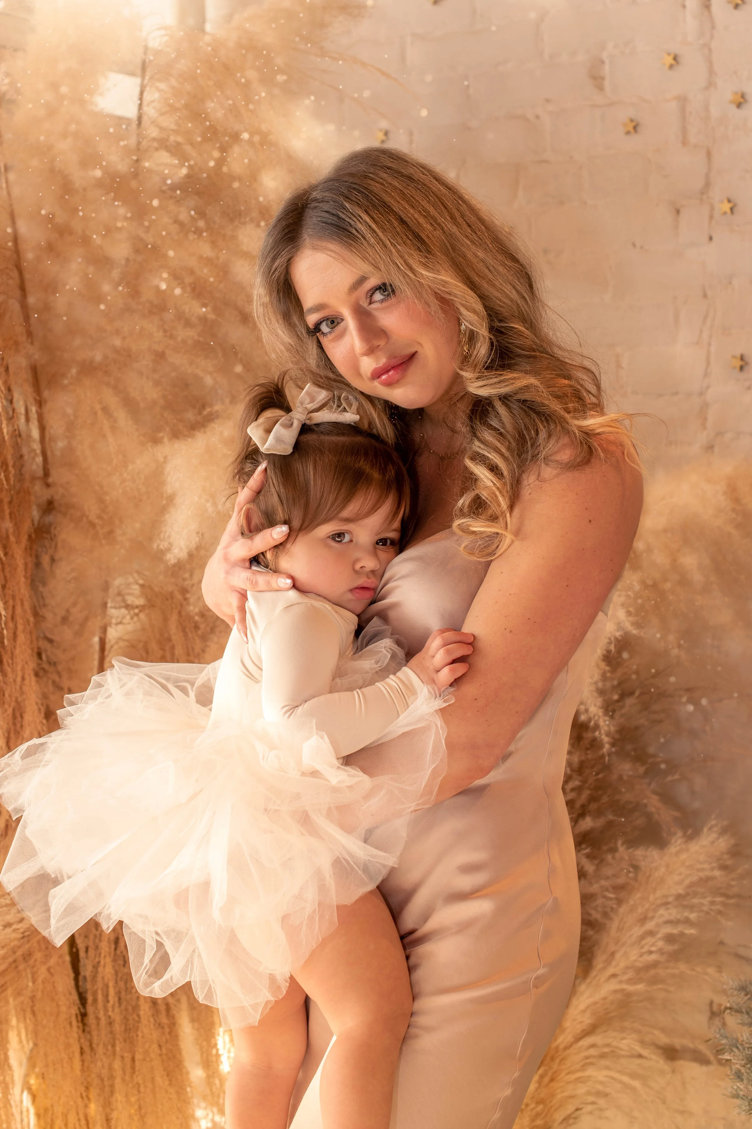 A woman holding a young girl dressed in a fancy cream-colored dress and bow, in a festive, winter-themed setting with golden decorations and stars on the wall.