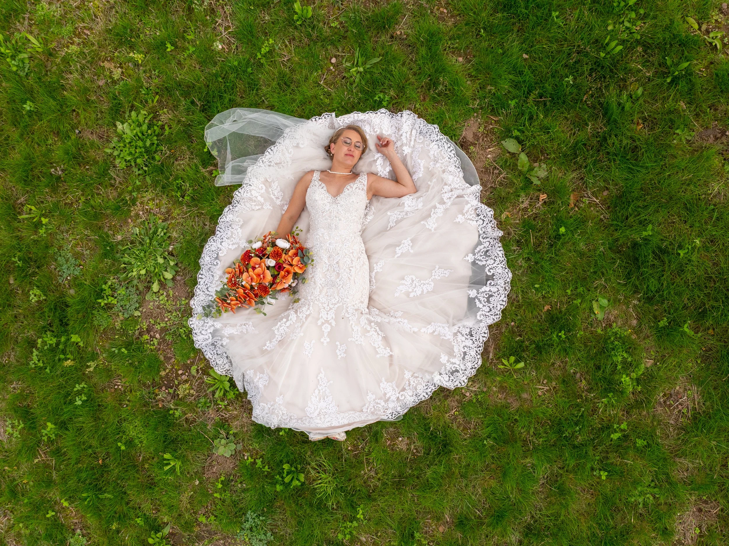 A woman in a wedding dress lying on a grassy field, holding a bouquet of orange and red flowers.